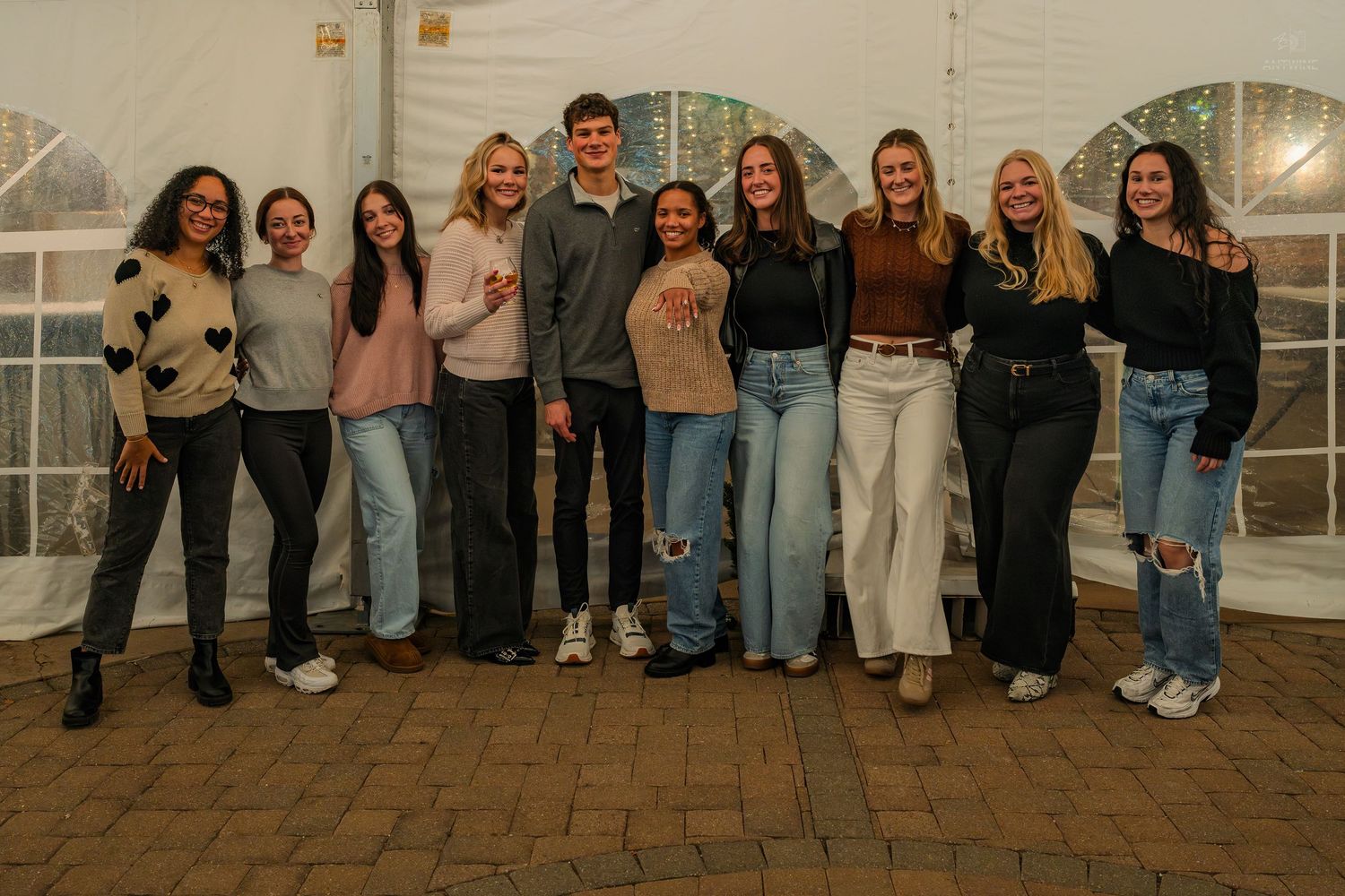 Group of ten young adults smile together in a line inside white event tent.