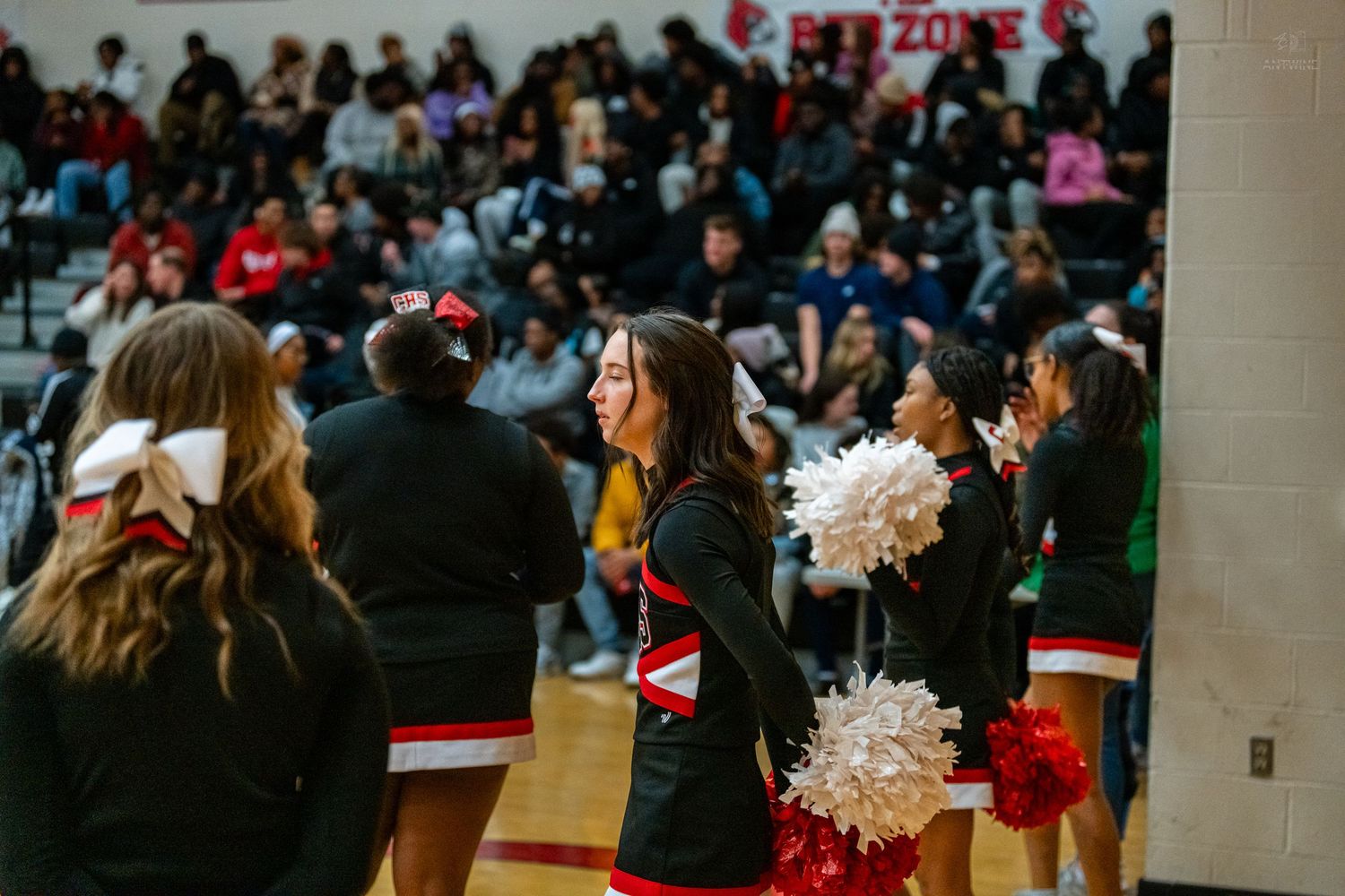 High school cheerleaders in red and black uniforms cheer with pom-poms at a basketball game.