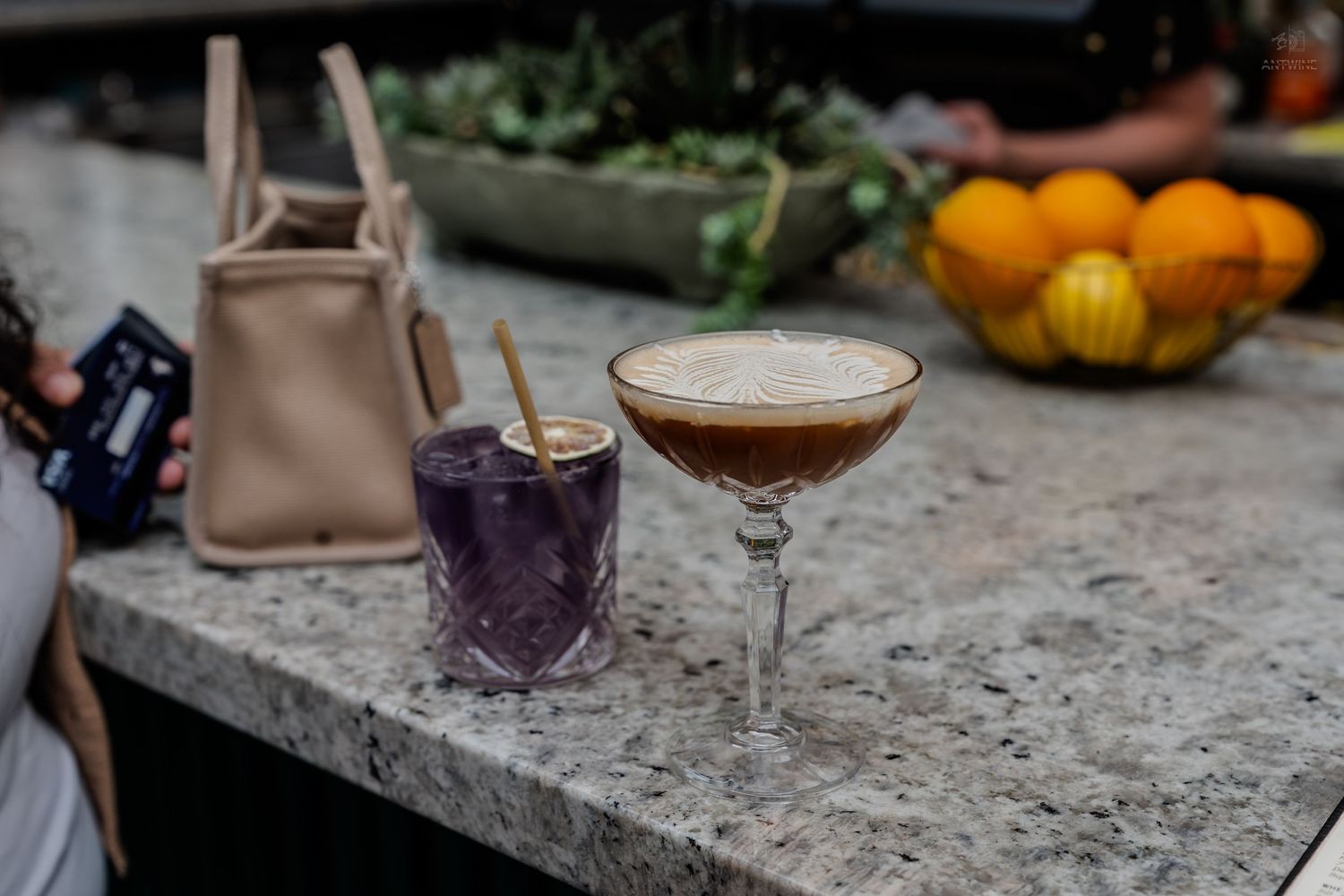 Two cocktails on marble bar top with handbag and fruit bowl in background.