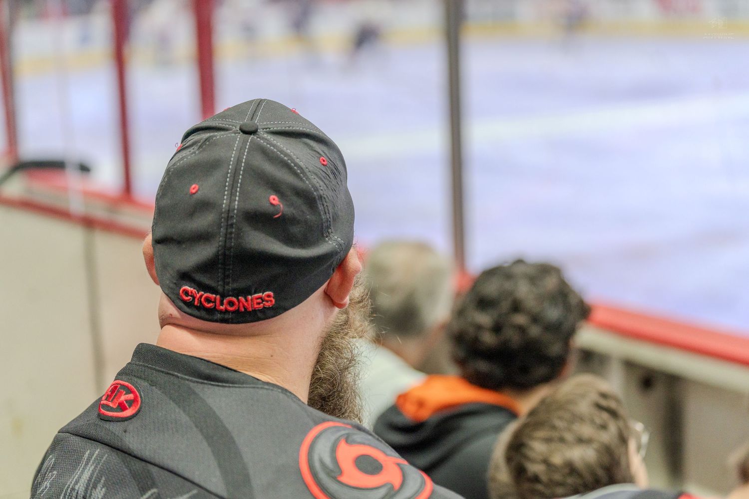 Cyclones fan wearing team cap watches ice hockey game from rinkside seats.