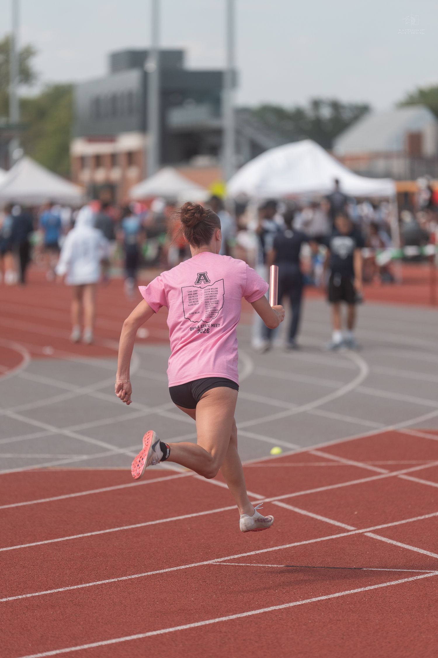 Female relay runner in pink shirt carries baton during outdoor track and field meet.