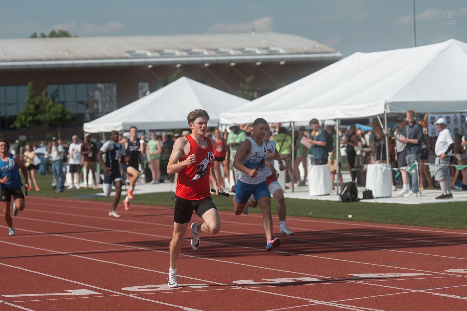 Runner in red jersey leads the race down the straightaway at an outdoor high school track meet.