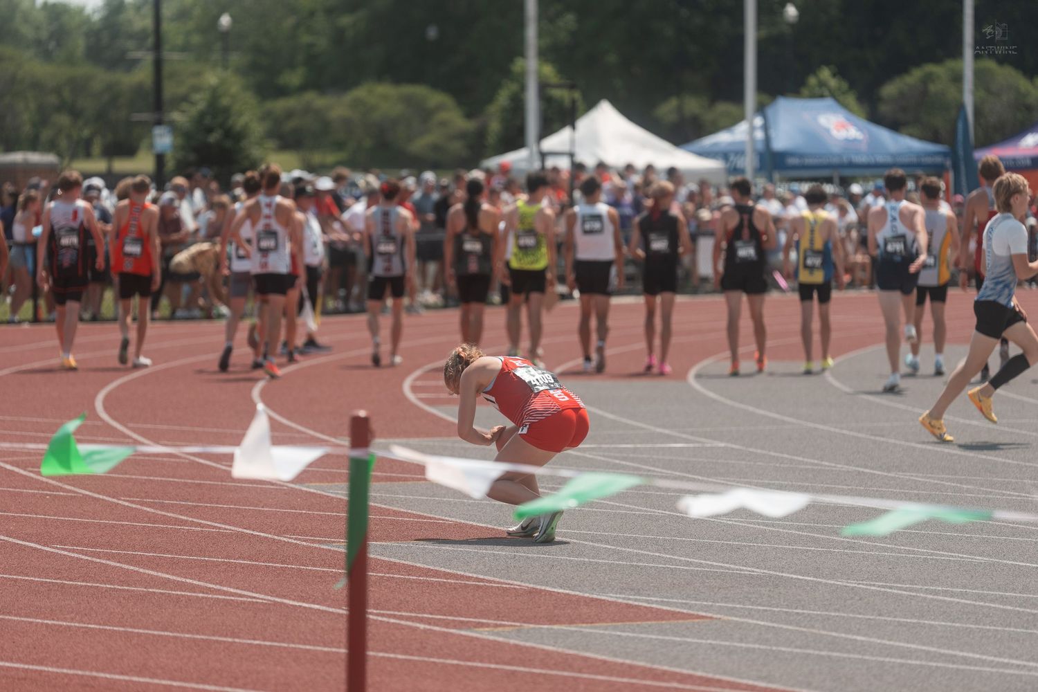 Tired athlete in red bends down on track after completing race at busy outdoor meet.