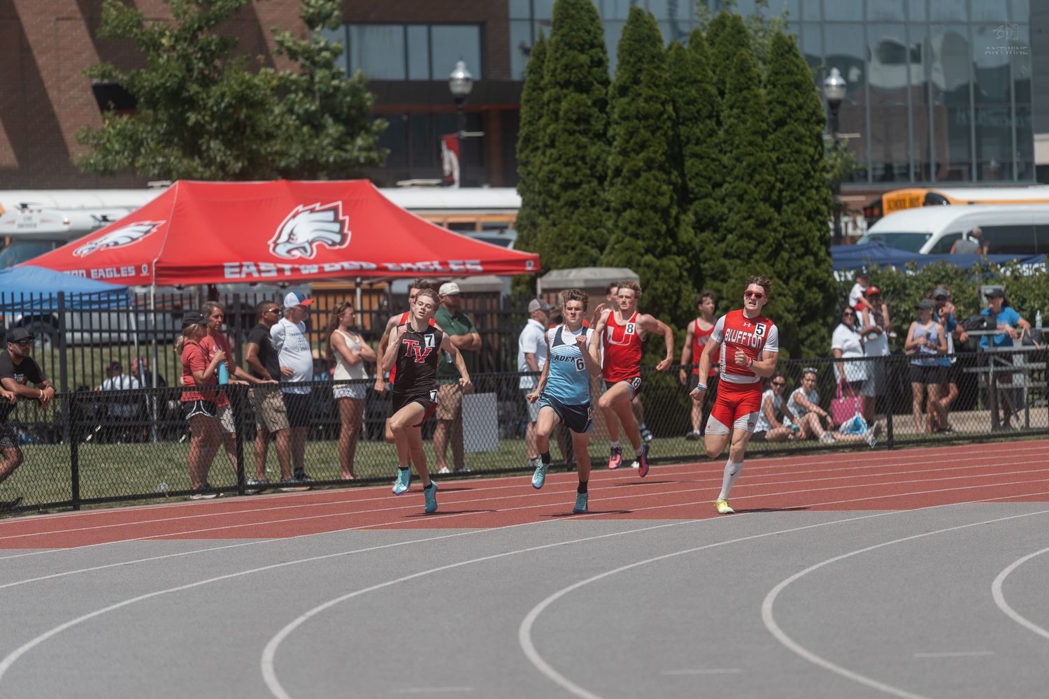 High school runners competing in a track race near an East tent at an outdoor athletic stadium.