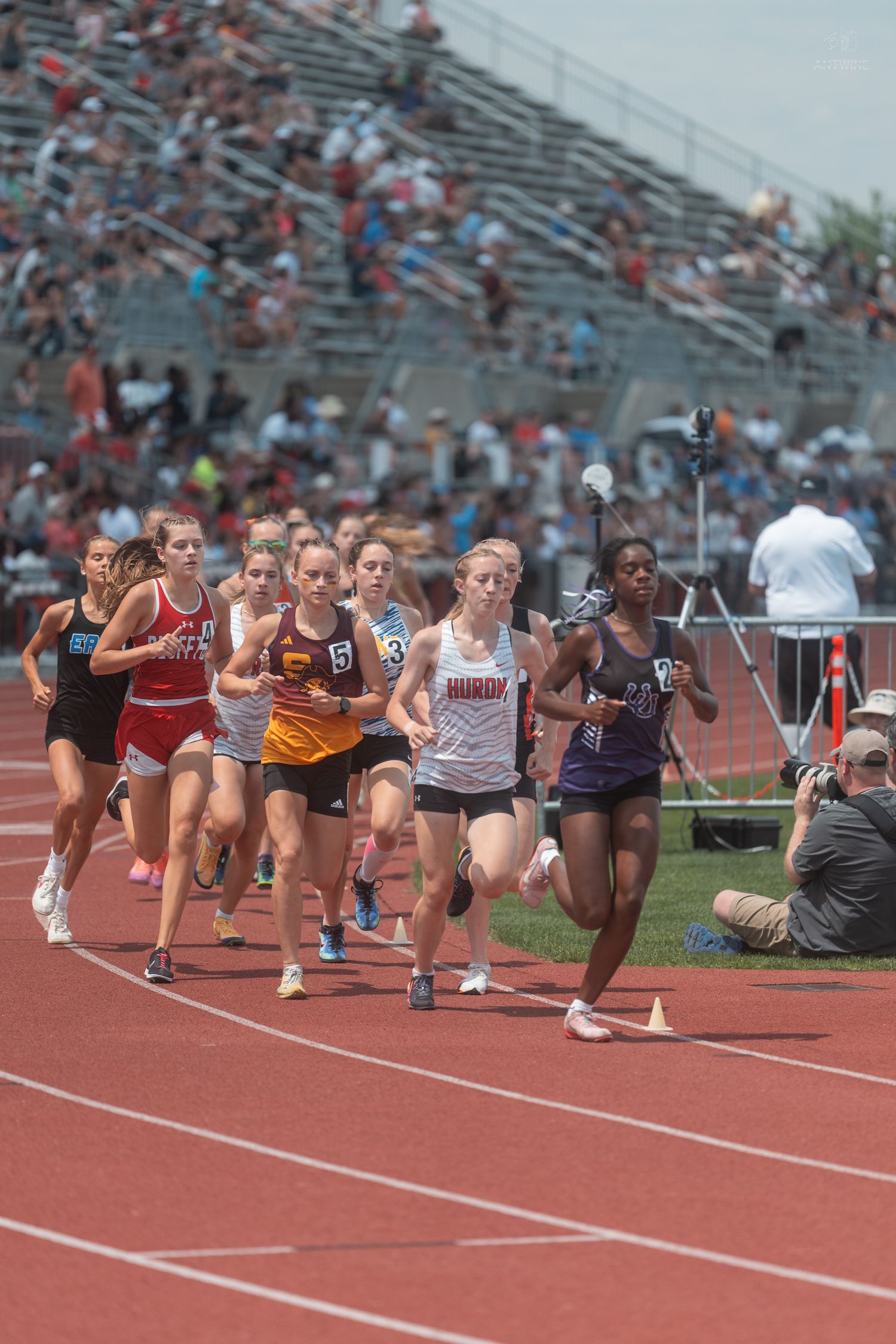 A large group of female athletes race on a red track at a competitive high school track meet.