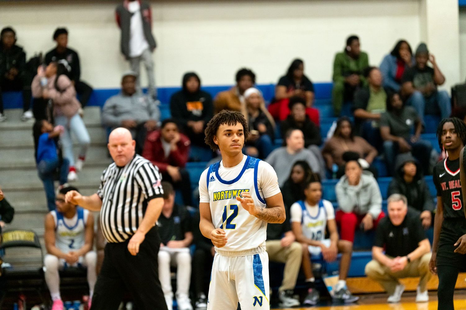 Northwest Knights player number 12 stands on court near referee during a high school basketball game with fans watching.