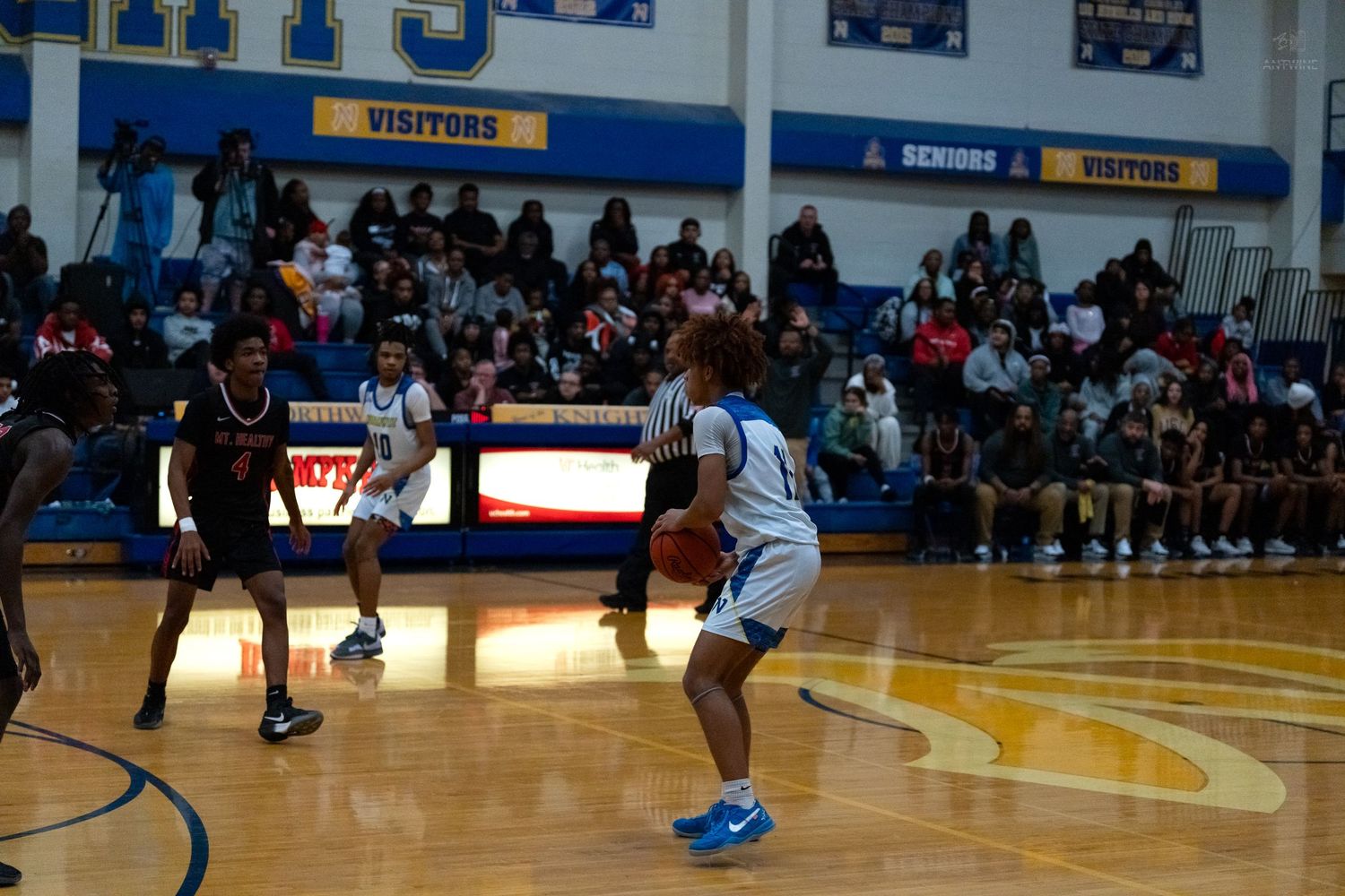High school basketball player in white uniform dribbles ball while defended by opponent in black during packed gym game.