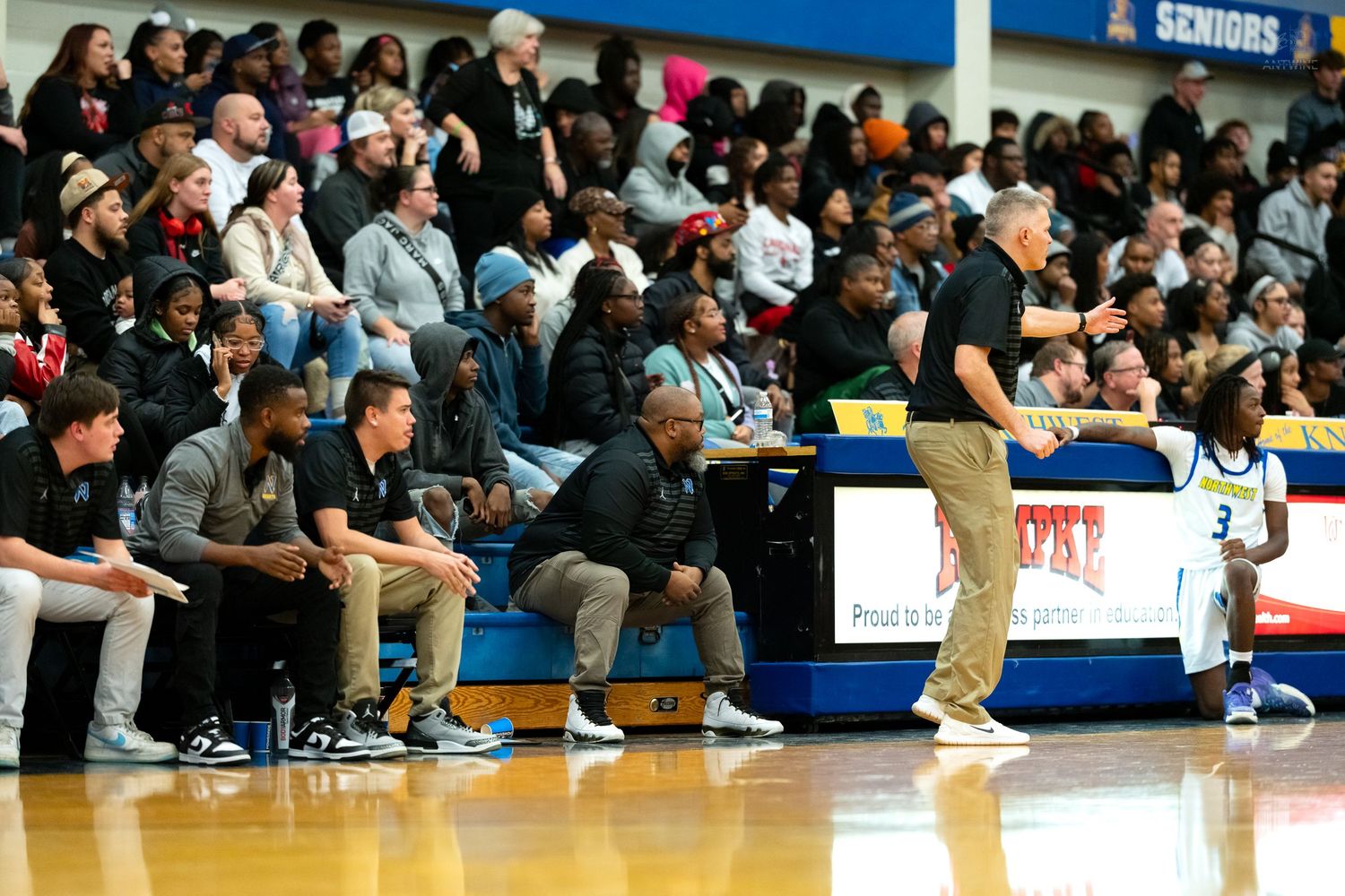 Basketball coaching staff watches game from sideline bench as player #3 stands nearby on the court.