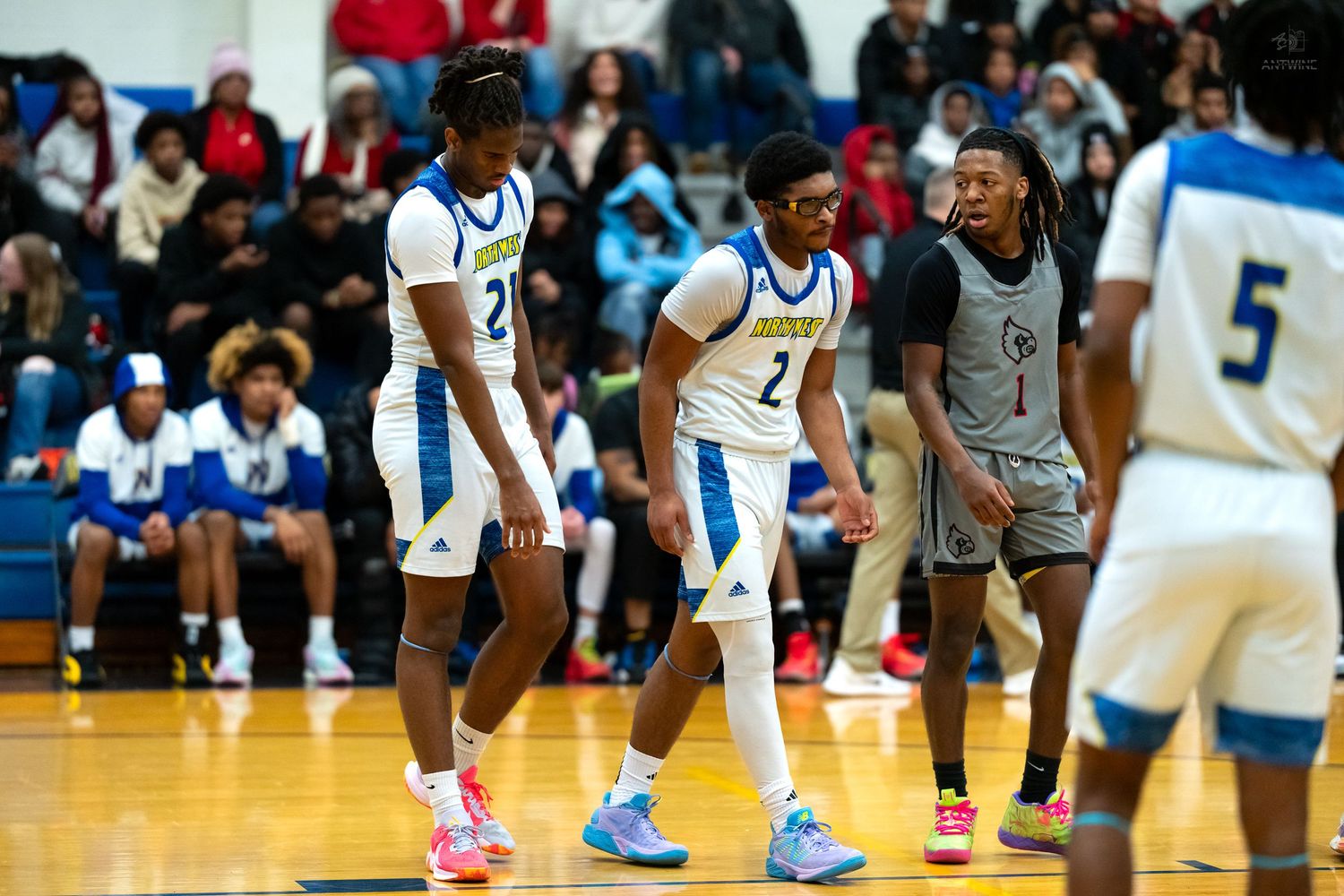 High school basketball players in white and blue uniforms face opponents on a crowded indoor court.