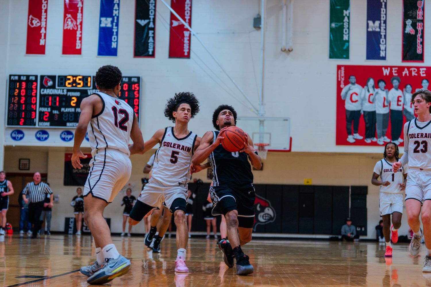 High school basketball player #0 drives to hoop past defenders with scoreboard visible in background.
