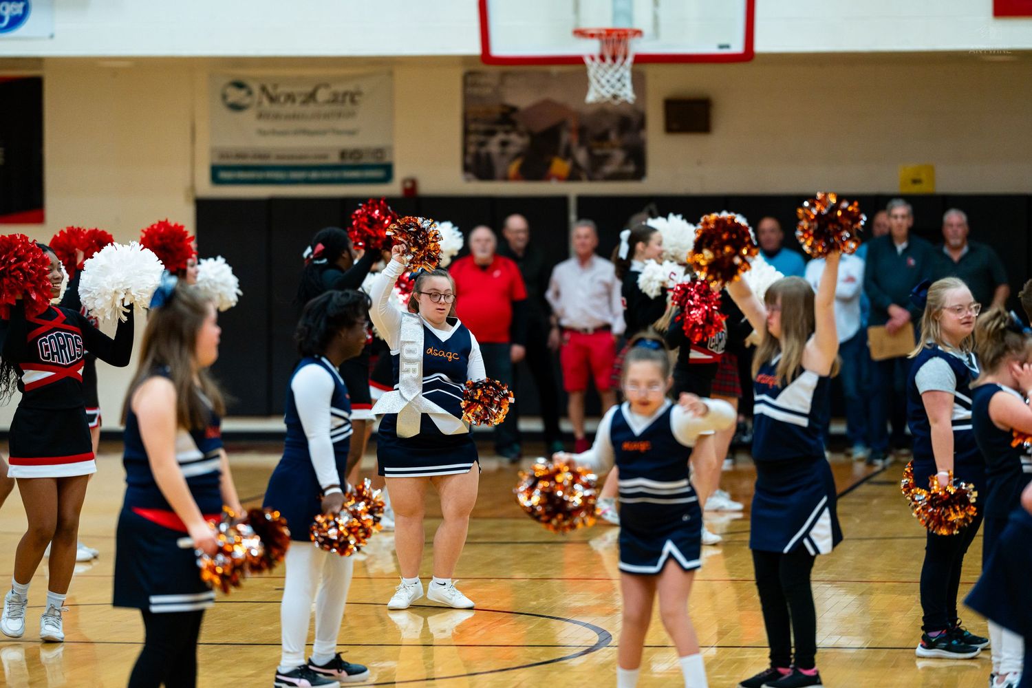 Cheerleaders in red and navy blue uniforms perform together with pom-poms in a school gymnasium.