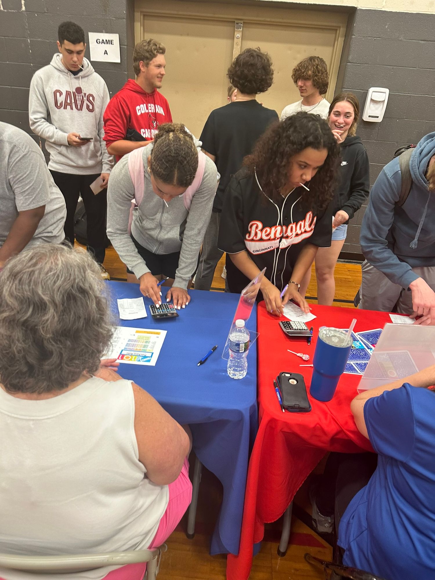 Students and community members gather around blue tables in a gym, signing papers and using phones at an event.