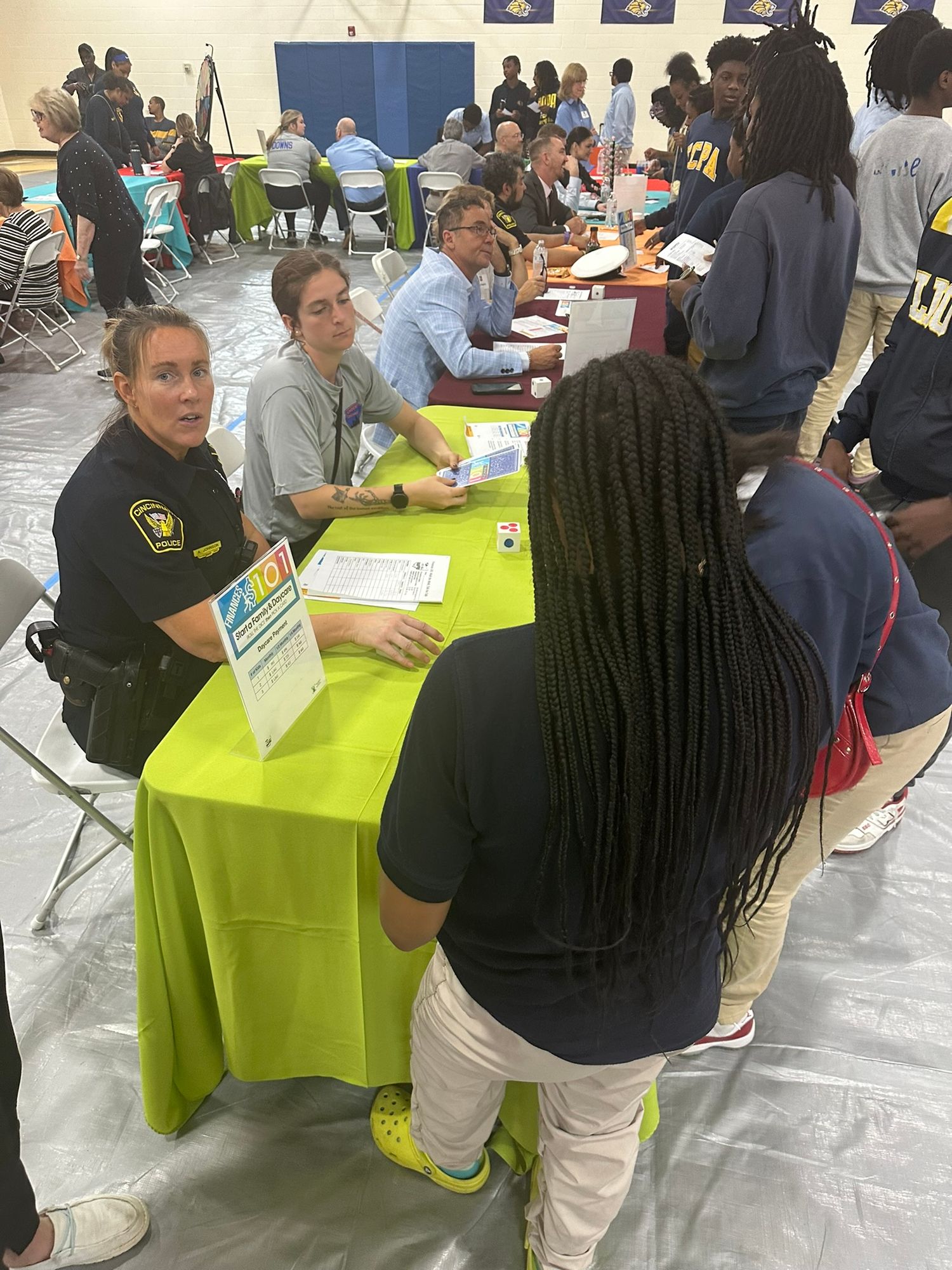 Students and officers engage at a community career fair event, seated at bright green tables in a school gymnasium.