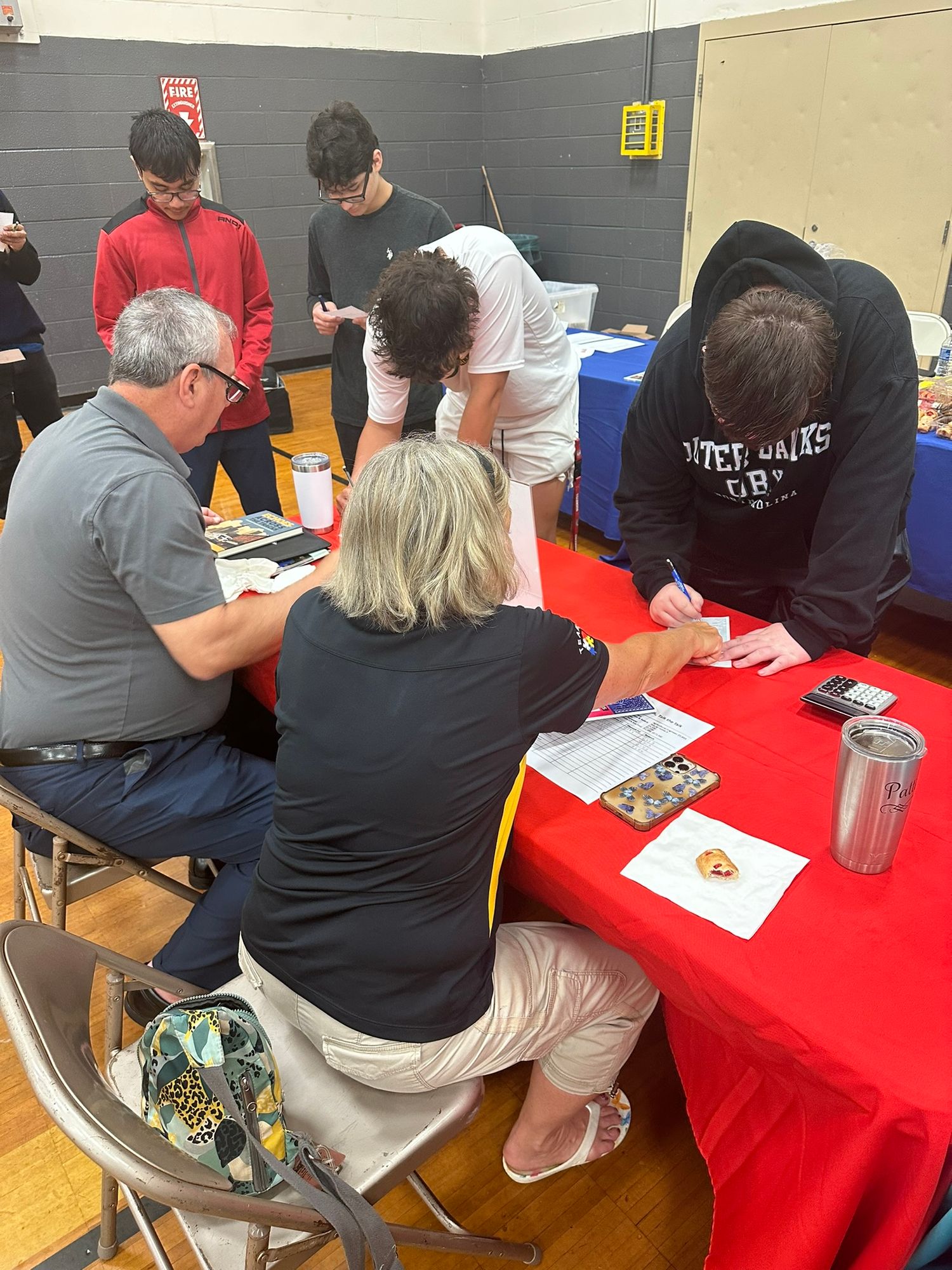 Adults and students gather around a red table at a school gymnasium event, reviewing and signing documents together.
