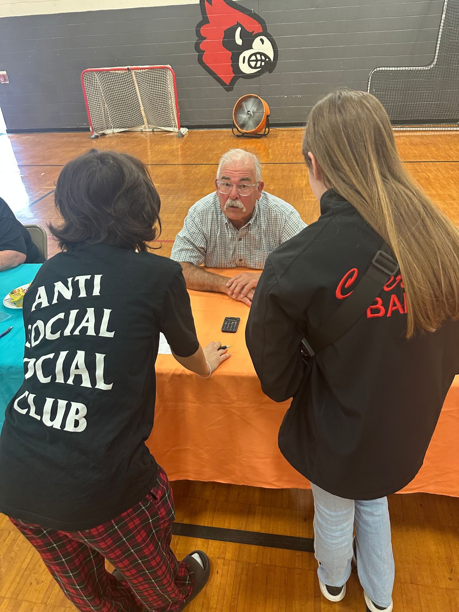 Students talk with an elderly man at a gym table during a school career or mentorship event.