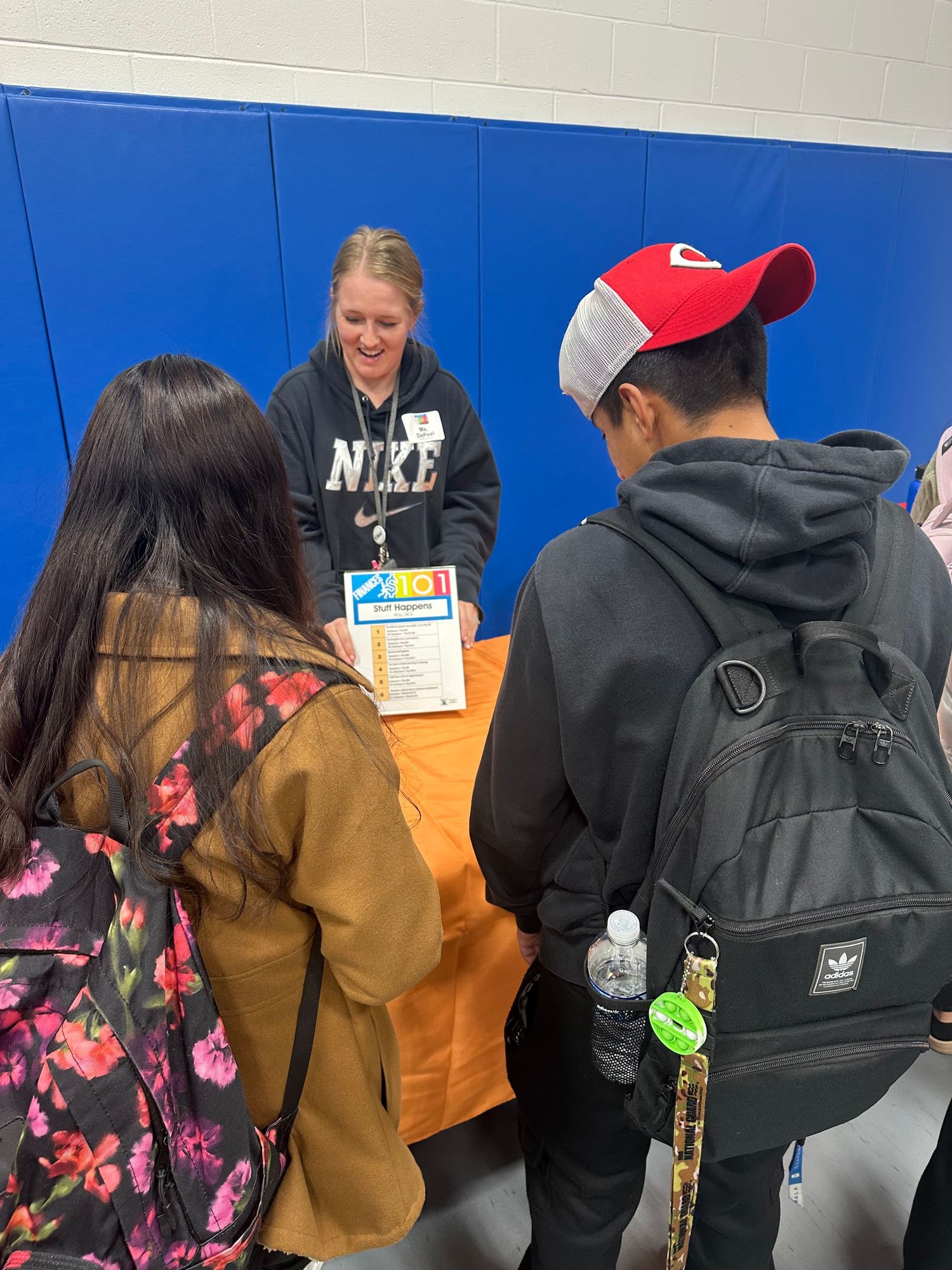 Students gather around an orange table speaking with a smiling Nike-clad representative at a school career fair in a gymnasium.