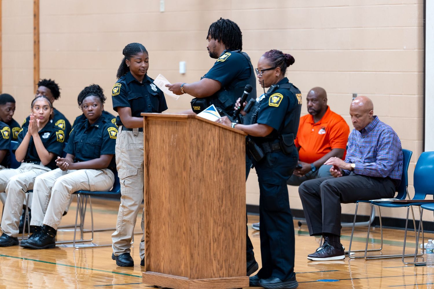Law enforcement officers present at a community meeting in a gymnasium with attendees seated in the background.