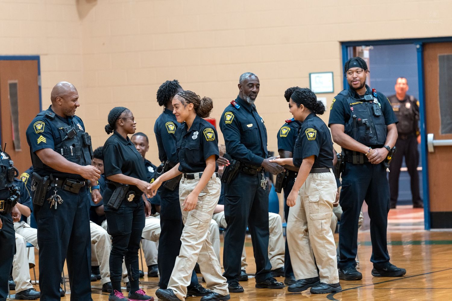 Black police officers and recruits in tan uniforms shake hands during a law enforcement graduation ceremony.