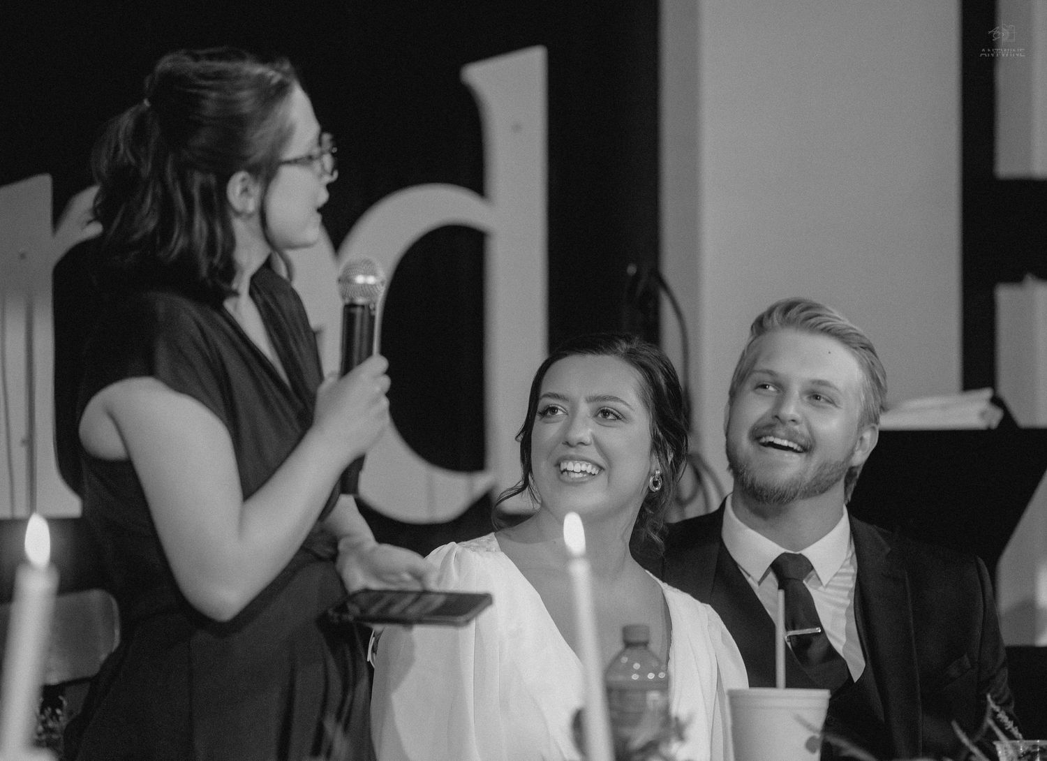 Black and white photo of a woman speaking into a microphone at a wedding reception with a smiling bride and groom seated nearby.