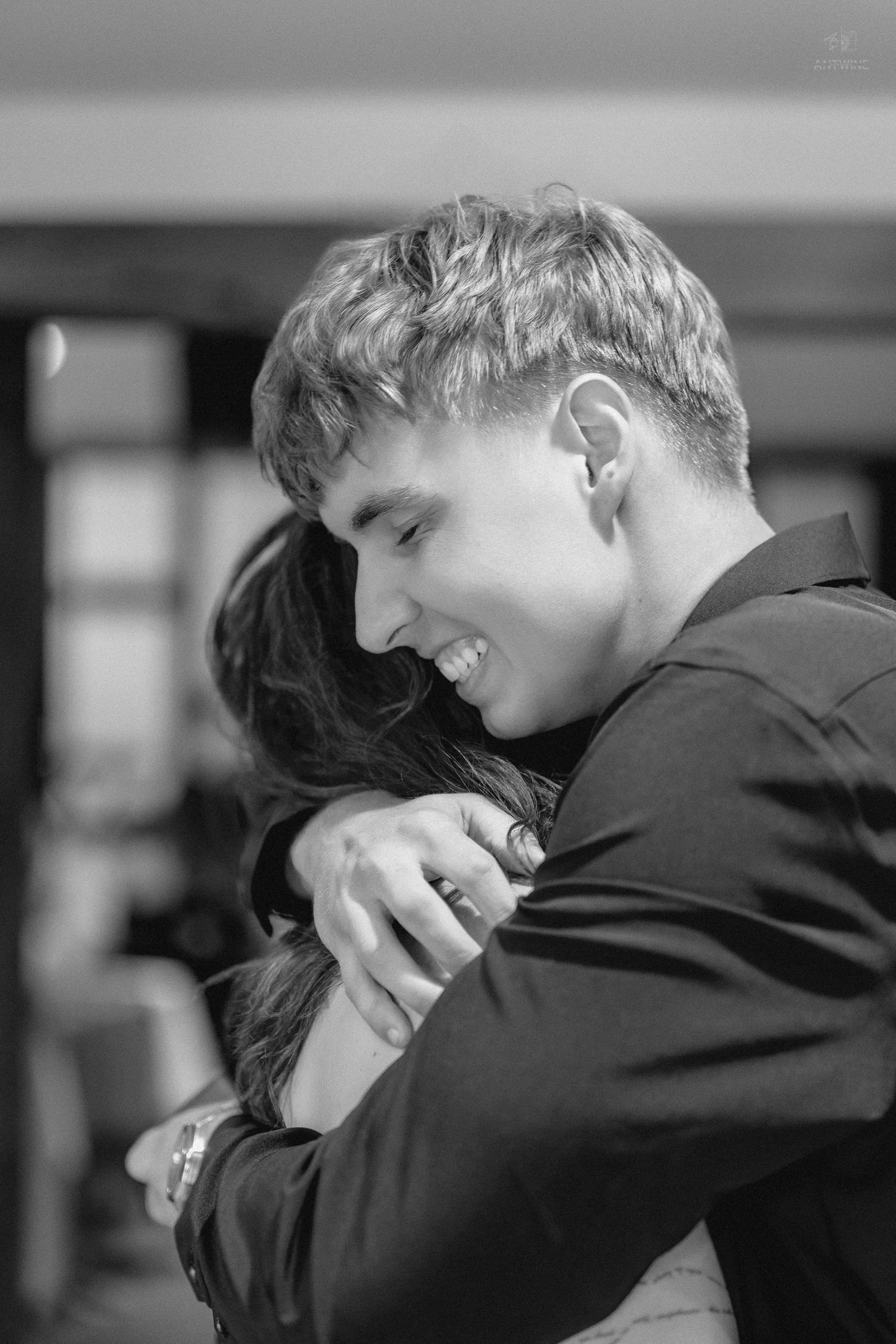 Smiling young man in black suit tenderly embraces someone at a wedding reception in black and white.