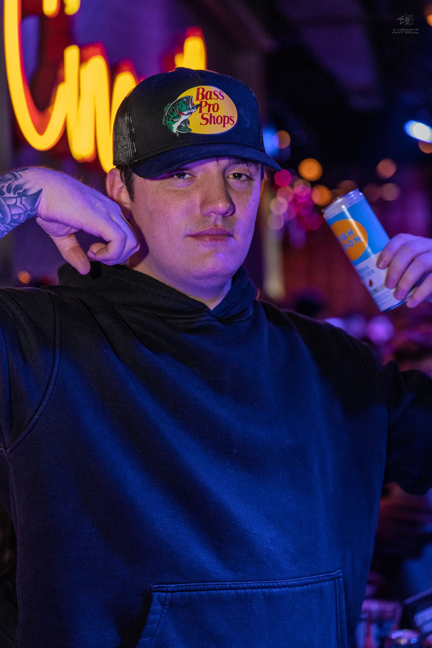 Young man wearing a Bass Pro Shops cap holding a drink can at a neon-lit bar or nightclub.