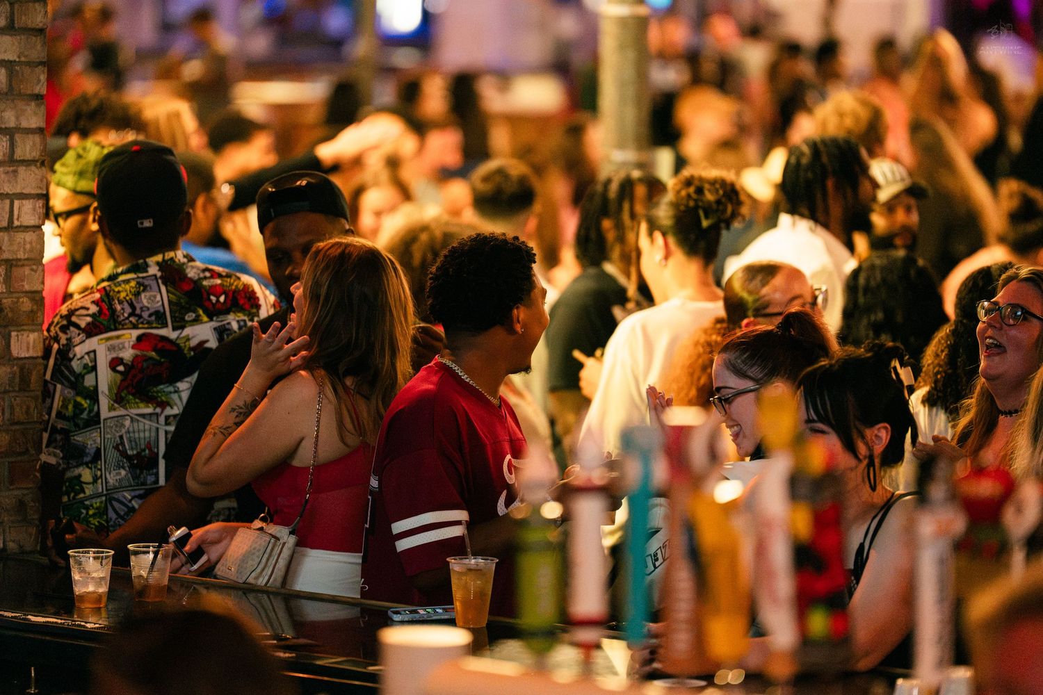 Large crowd of young adults socializing at a busy bar with drinks on the counter.