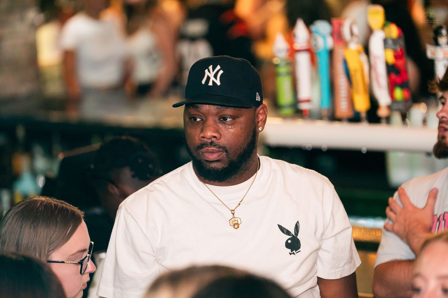 Man wearing a New York Yankees cap and Playboy white t-shirt standing at a busy bar.