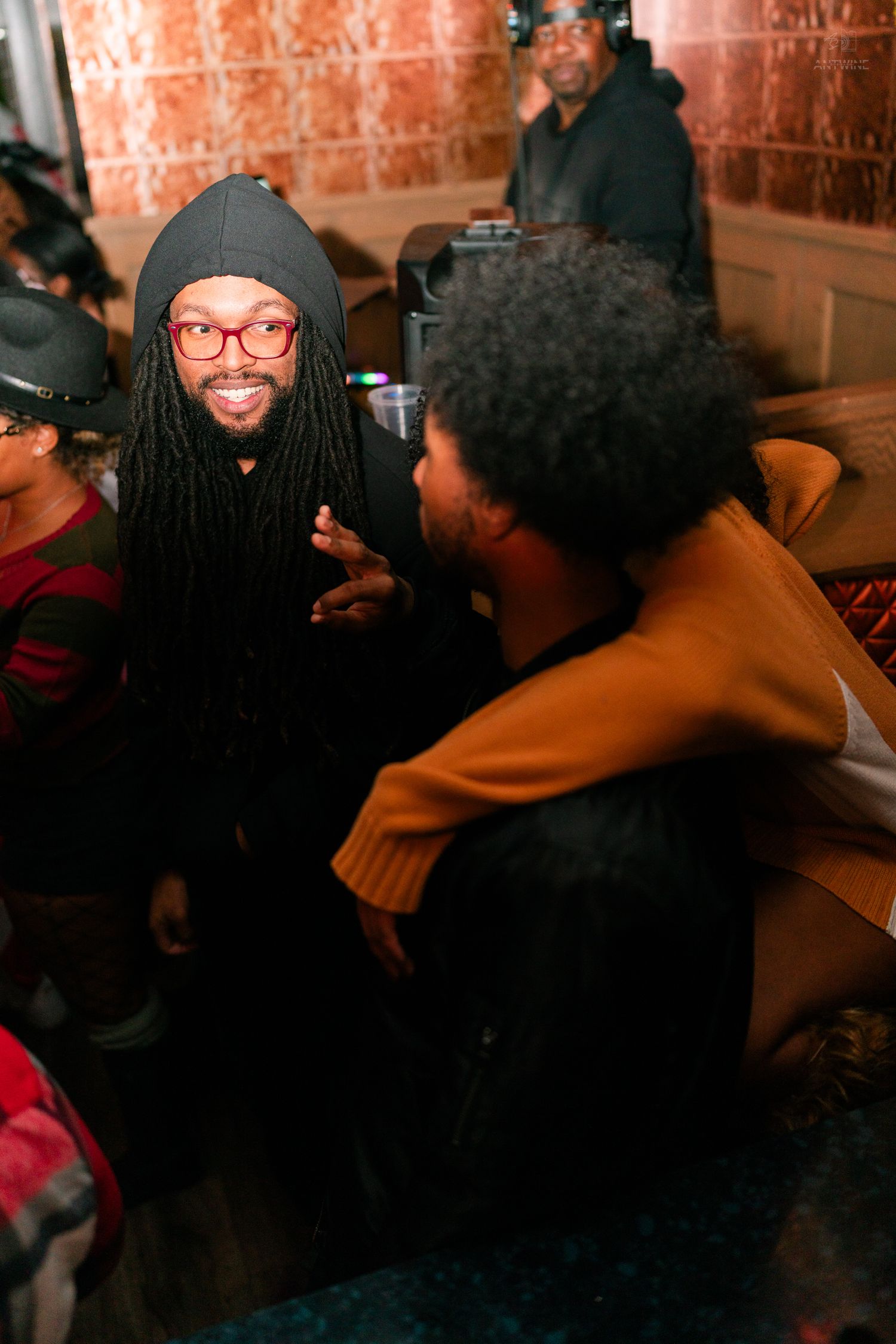 Man with dreadlocks and red glasses smiling while chatting with a friend at an indoor party.