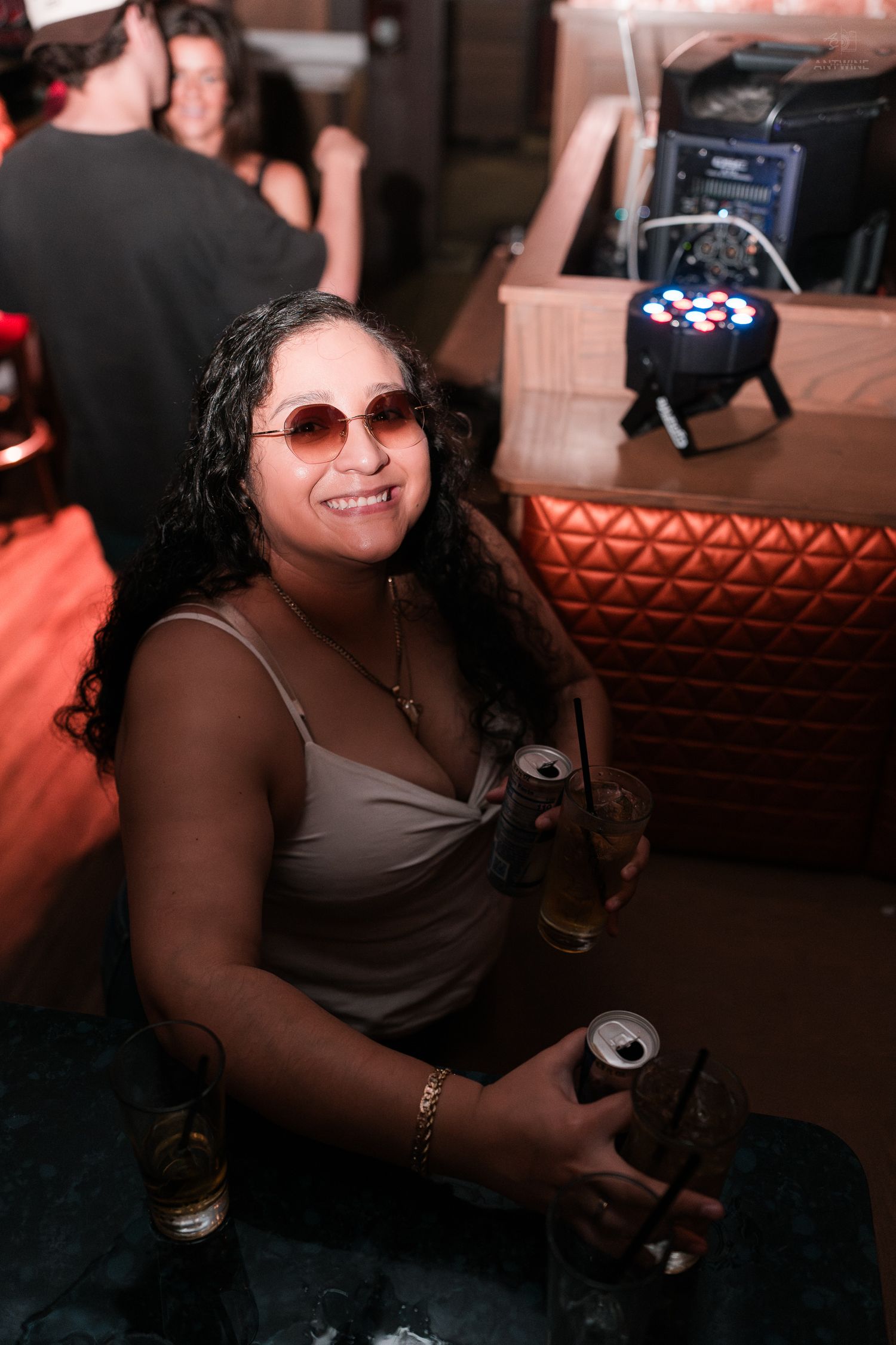 Woman with curly hair and round sunglasses smiling while holding drinks at a nightclub event.