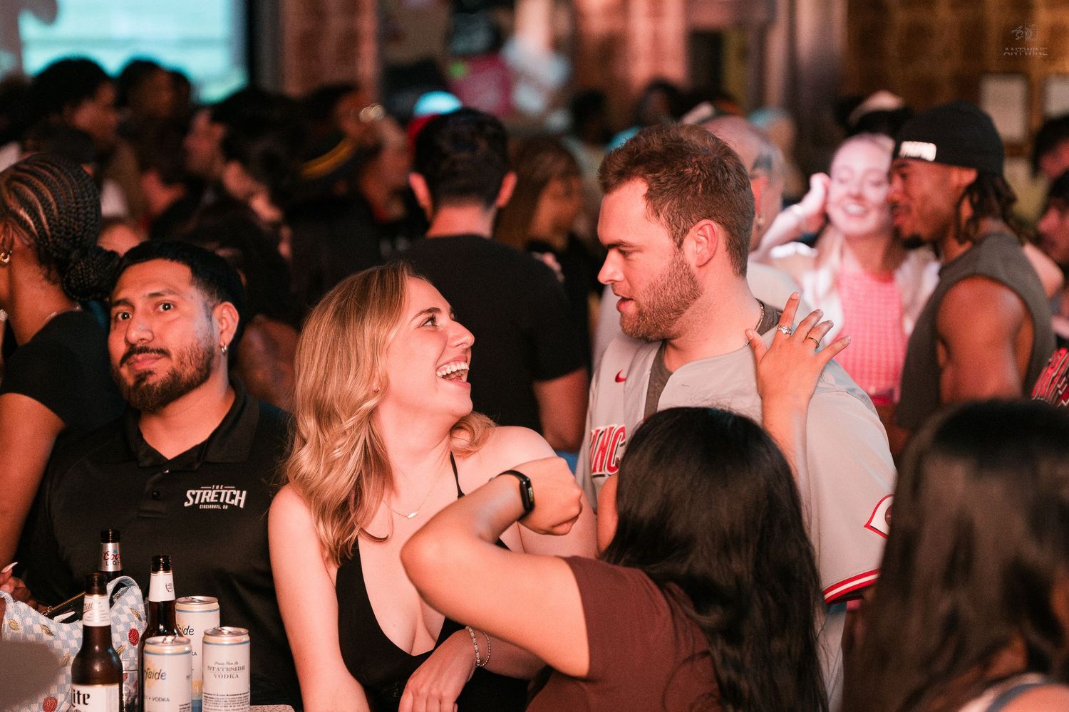 Couple smiling and chatting at a crowded nightclub bar with drinks on table.