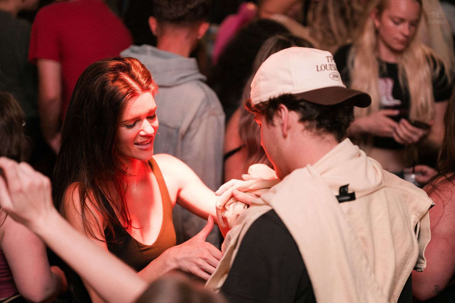Two people sharing an intimate conversation under warm red lighting at a crowded nightclub.