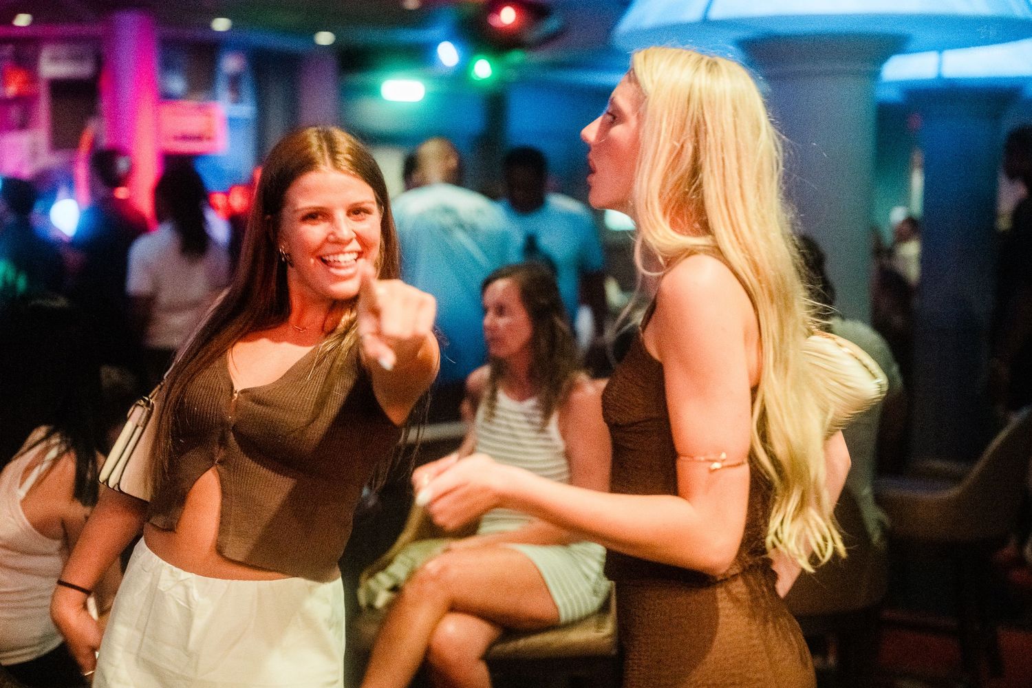Two women smile and dance together under colorful lights at a nightclub party.