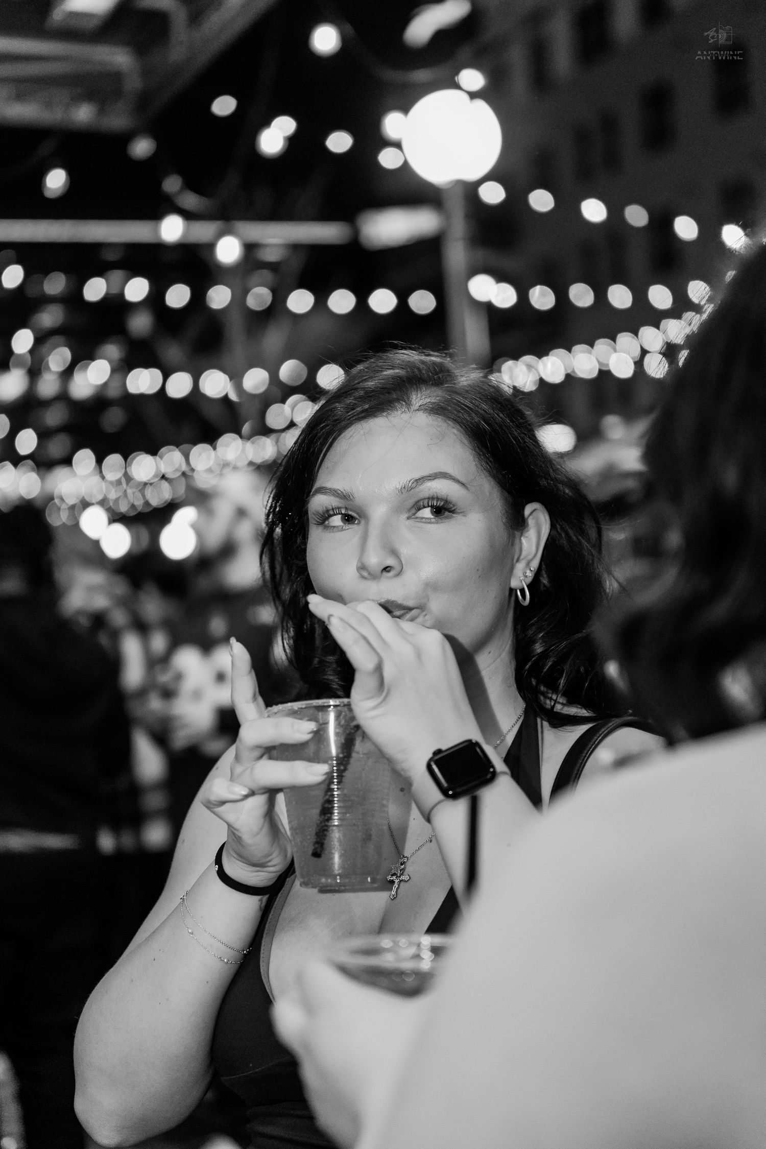 Black and white photo of a woman sipping a cocktail under string lights at a bar.