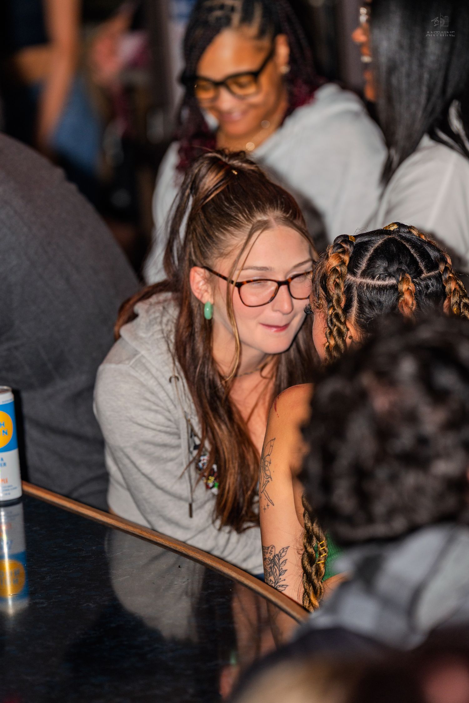 Young woman with long brown hair and glasses sits at a bar counter at a social event.