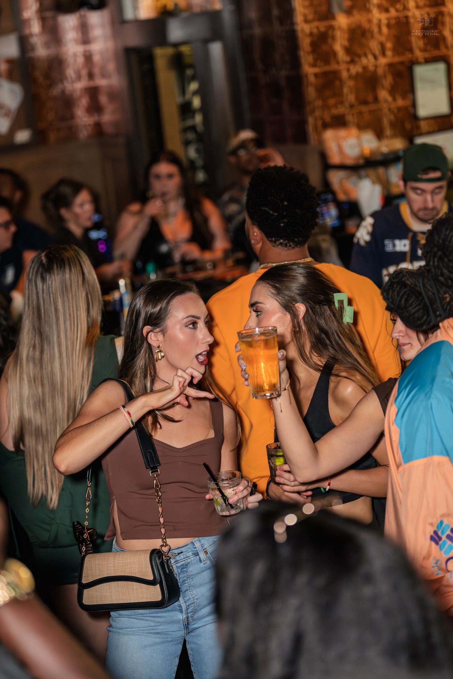 Two women in brown tops laughing and holding drinks at bar.
