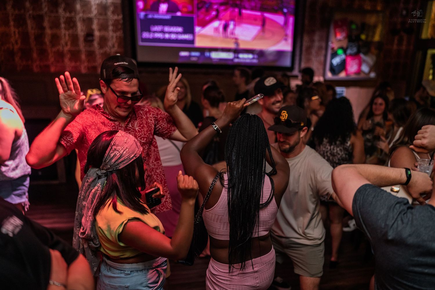 Crowd of people dancing inside a lively bar with a large sports screen and warm ambient lighting.