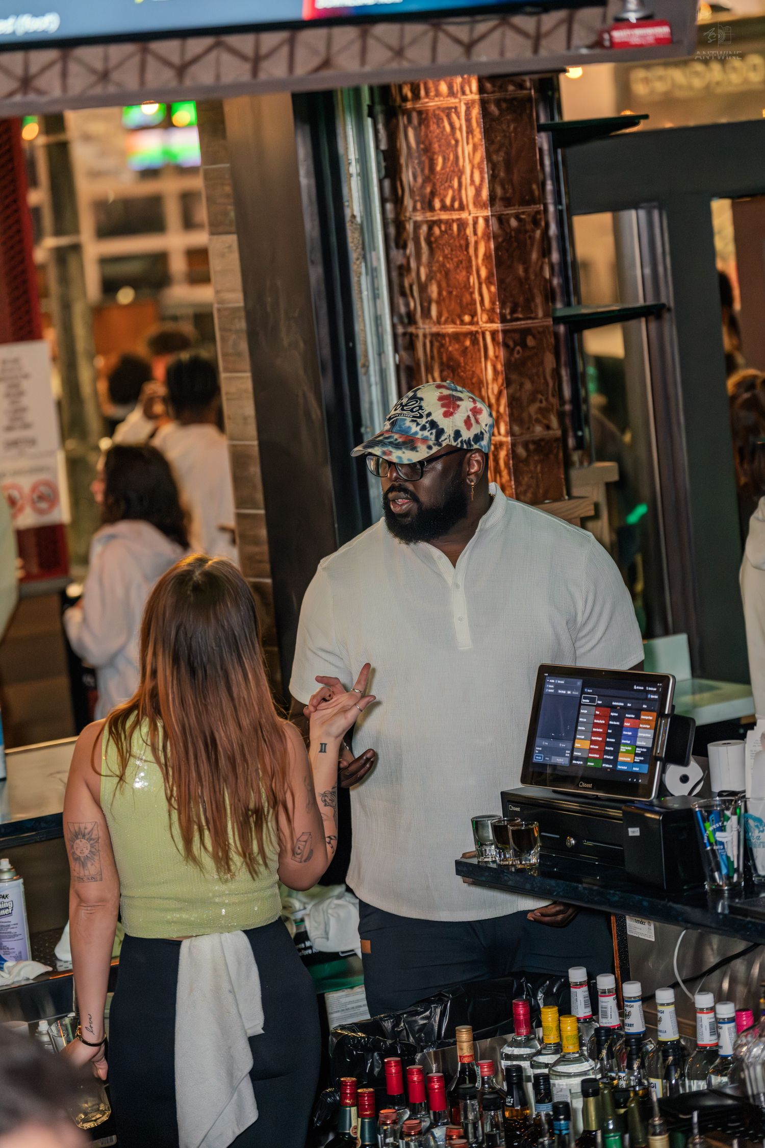 Bartender in a white shirt and patterned cap speaking with a customer at a busy bar.