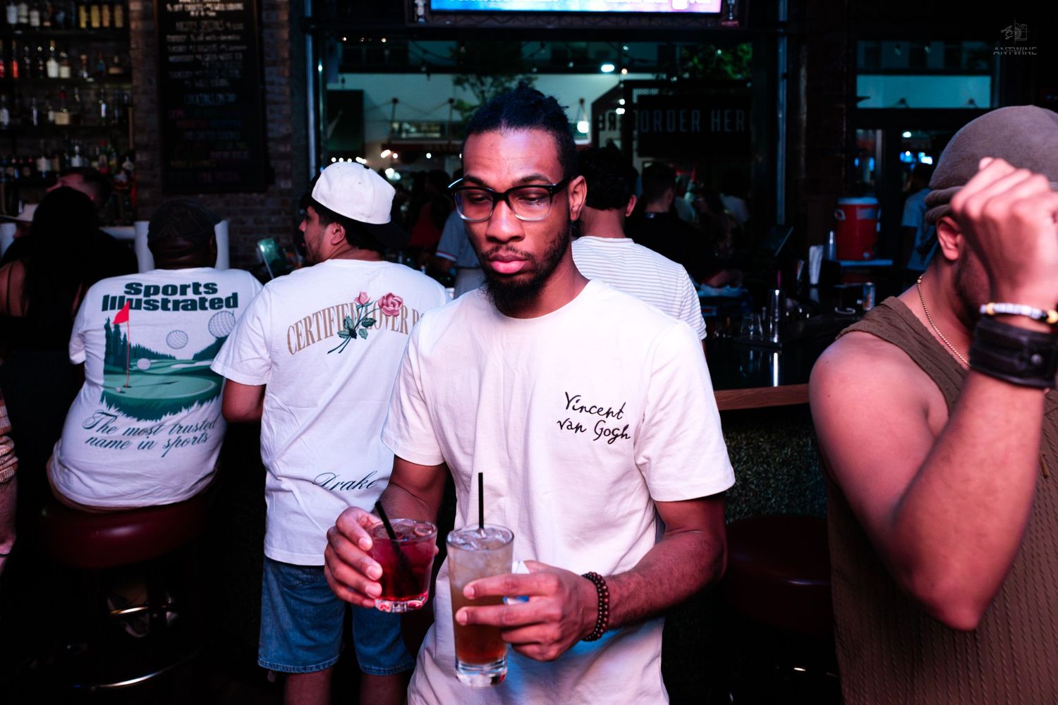 Young man in white shirt holding cocktails and posing confidently at a crowded indoor bar at night.