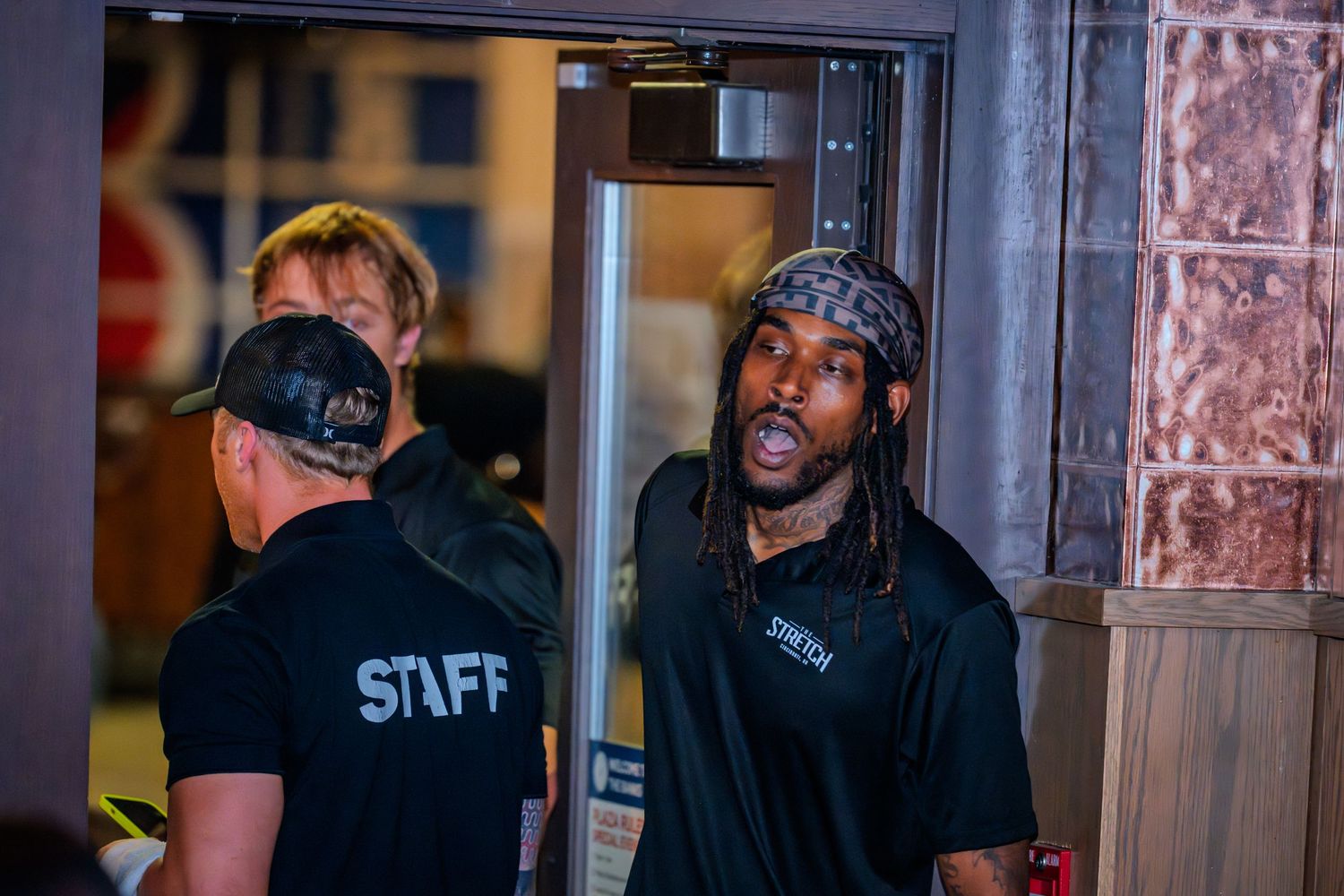 A man with dreadlocks in a black shirt speaks with staff members at a venue entrance.