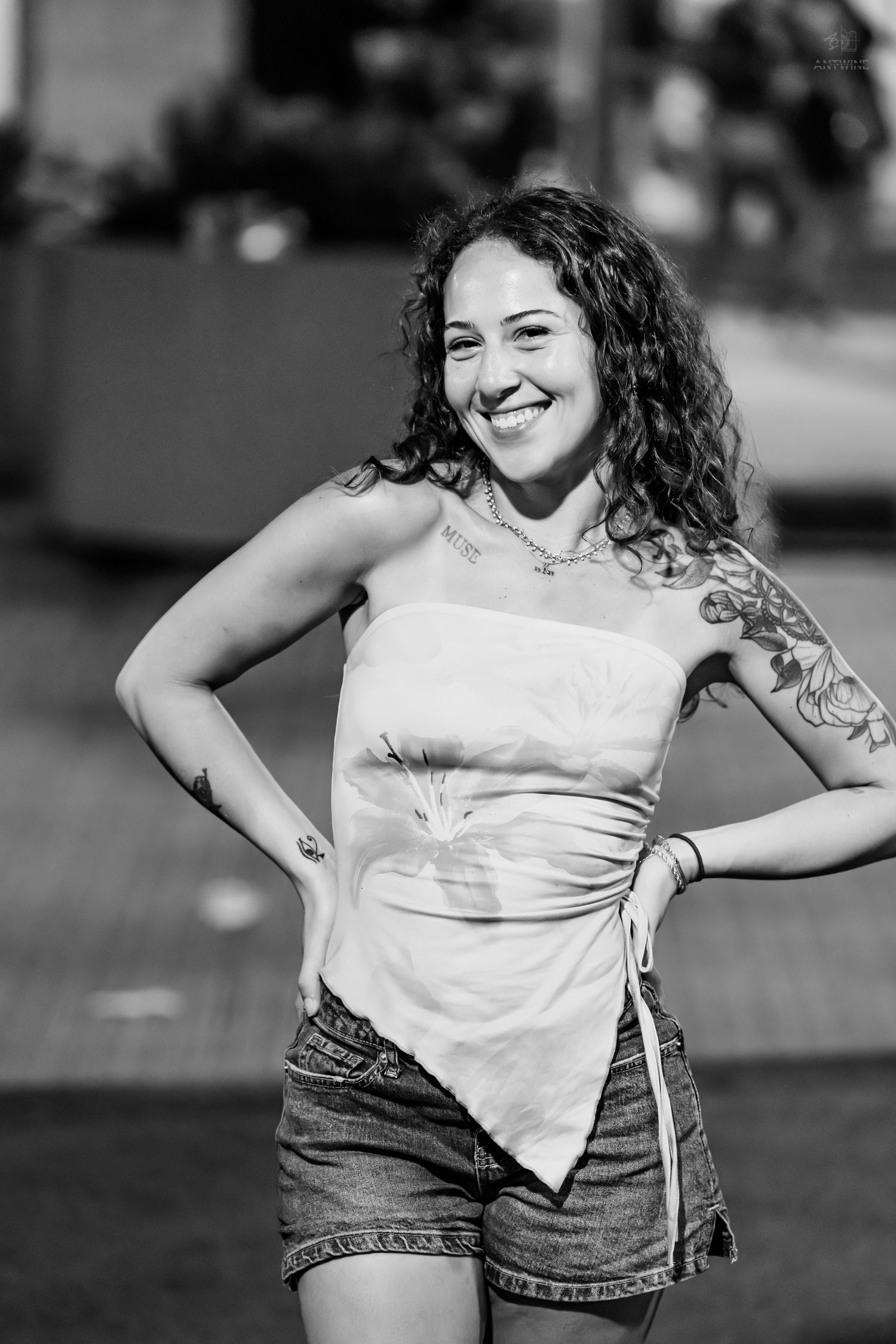A smiling tattooed woman in a strapless top and denim shorts poses with hands on hips in black and white.
