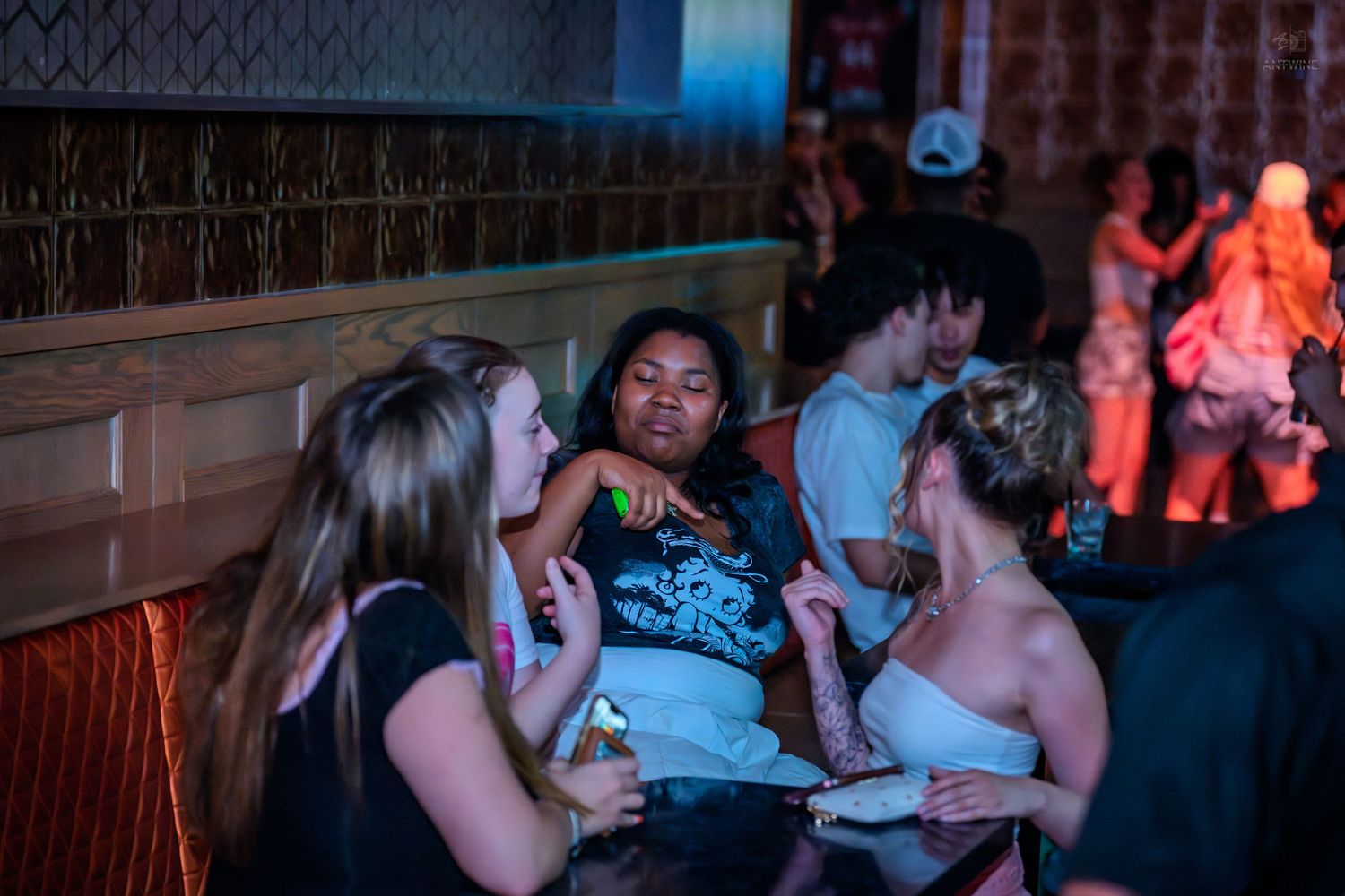 Three women chat and laugh together in a dimly lit, lively bar or nightclub setting.