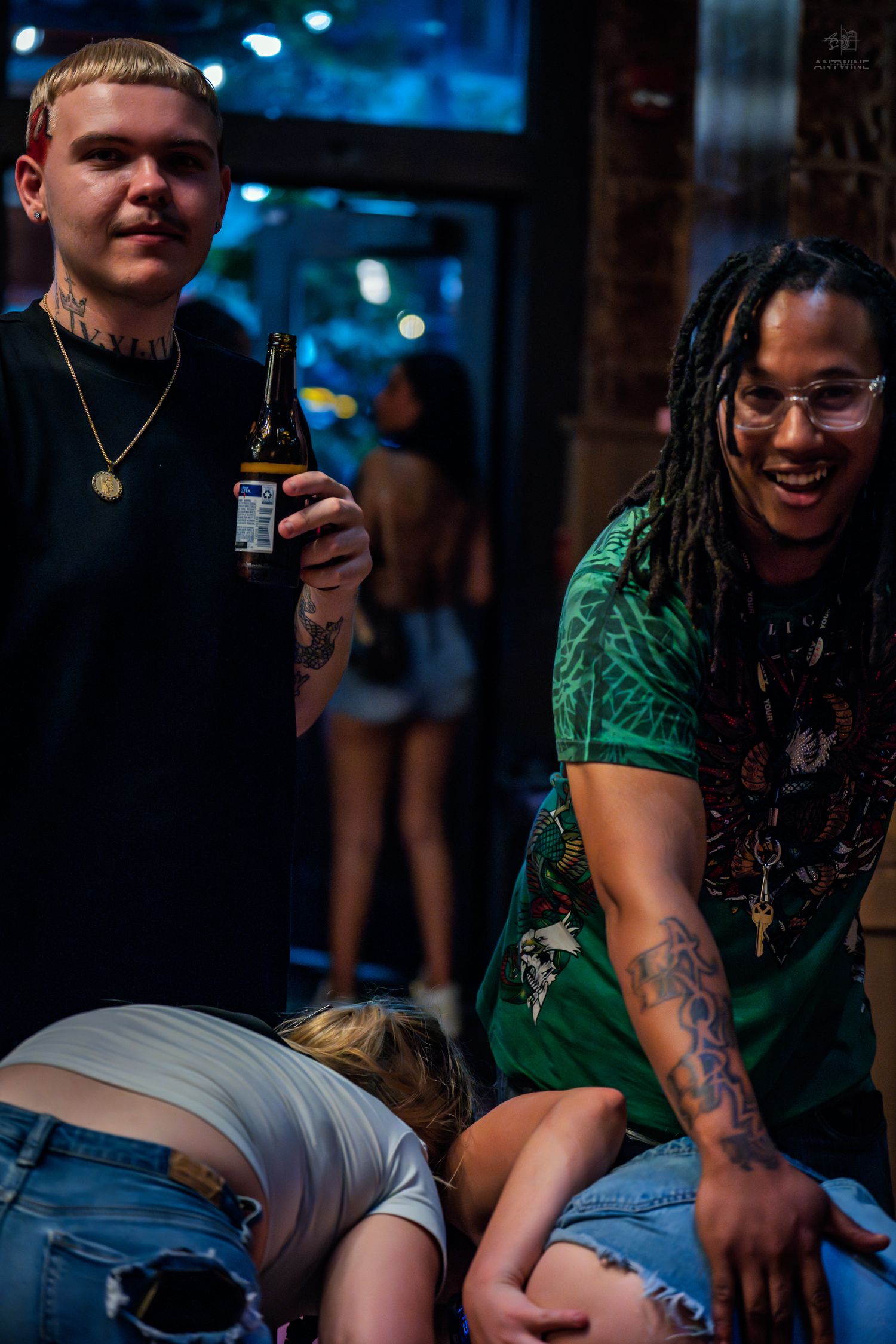 Two tattooed men smiling at a nightclub bar while a woman leans forward between them.