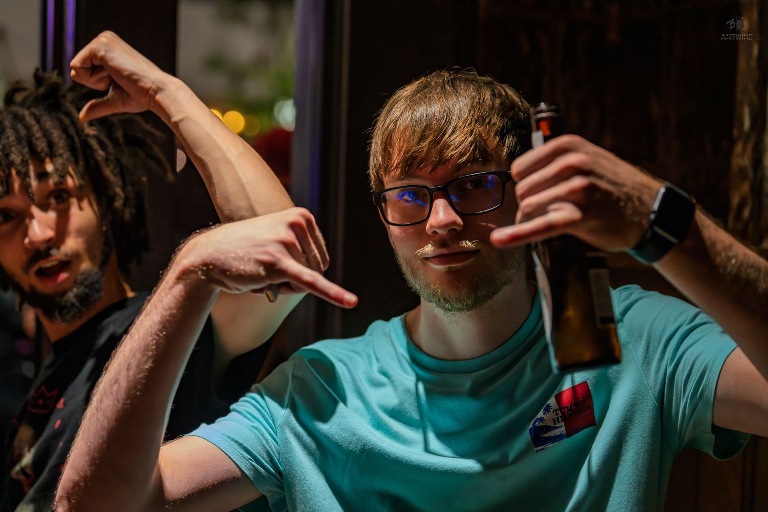 Young man in teal shirt holding a beer bottle and pointing at a bar.