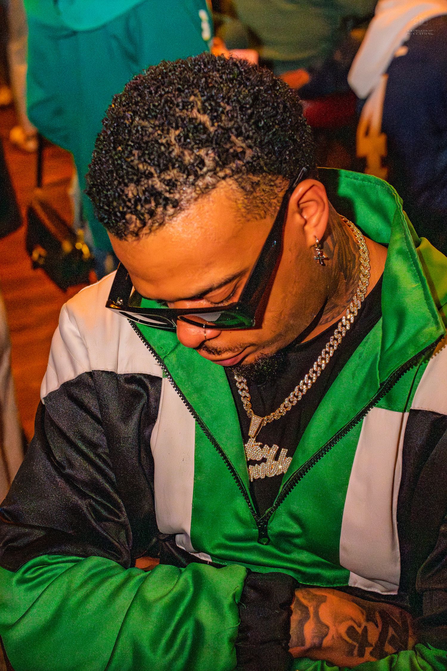 Man in green track jacket with gold chain jewelry and sunglasses looking down at a party event.