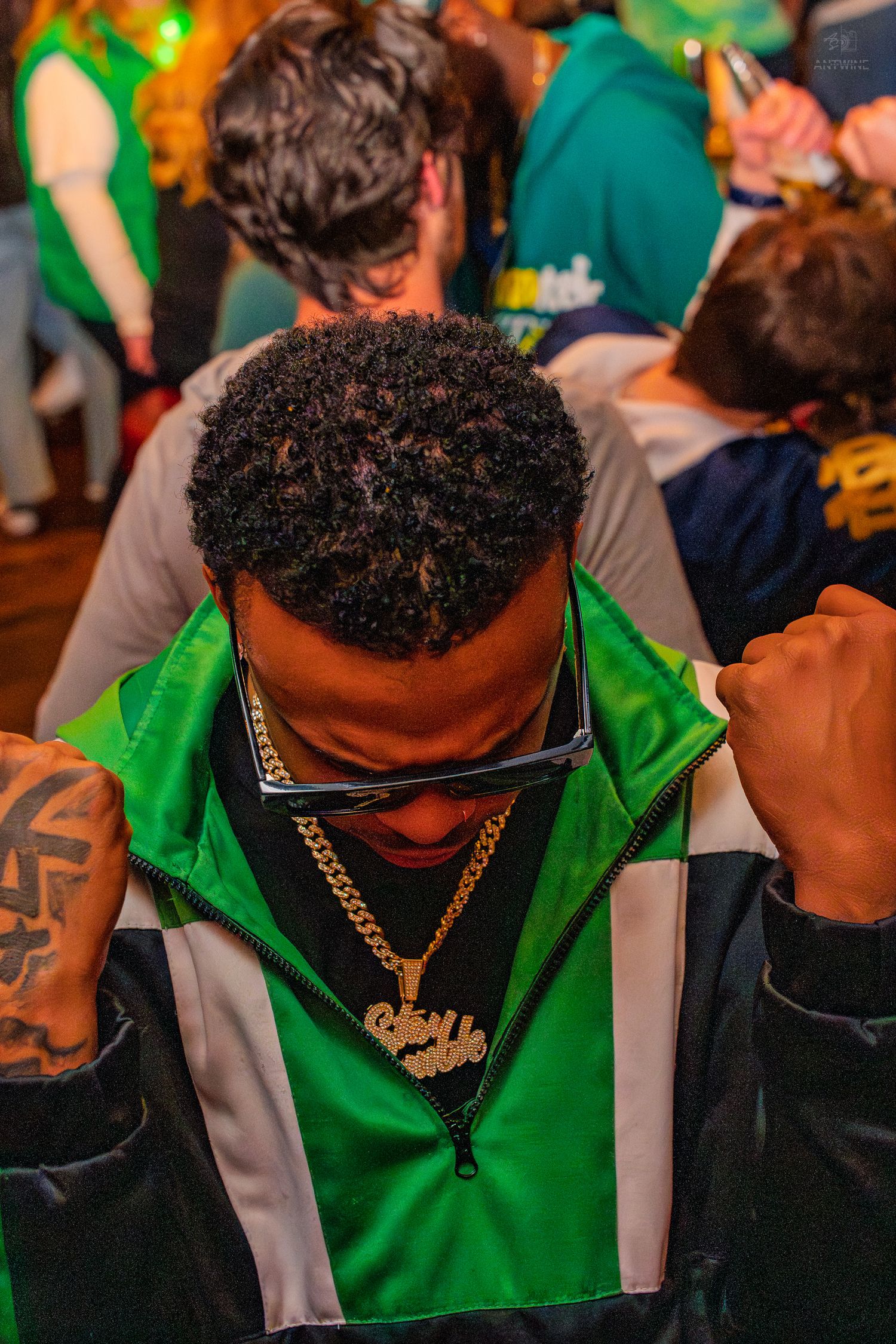Man in green jacket wearing gold chain necklace at a crowded St. Patrick's Day party.