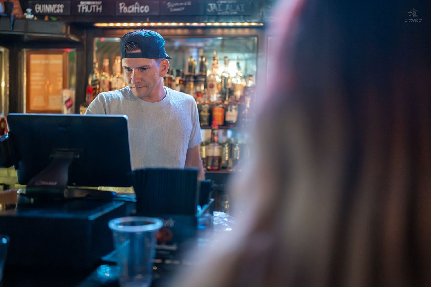 Bartender in a cap works behind a bar counter with liquor bottles and a POS system in a dimly lit bar.