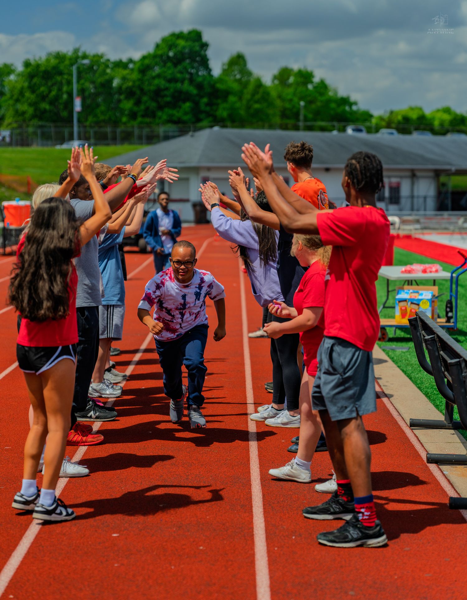Students and athletes form a celebratory high-five tunnel on a red running track outdoors on a cloudy day.