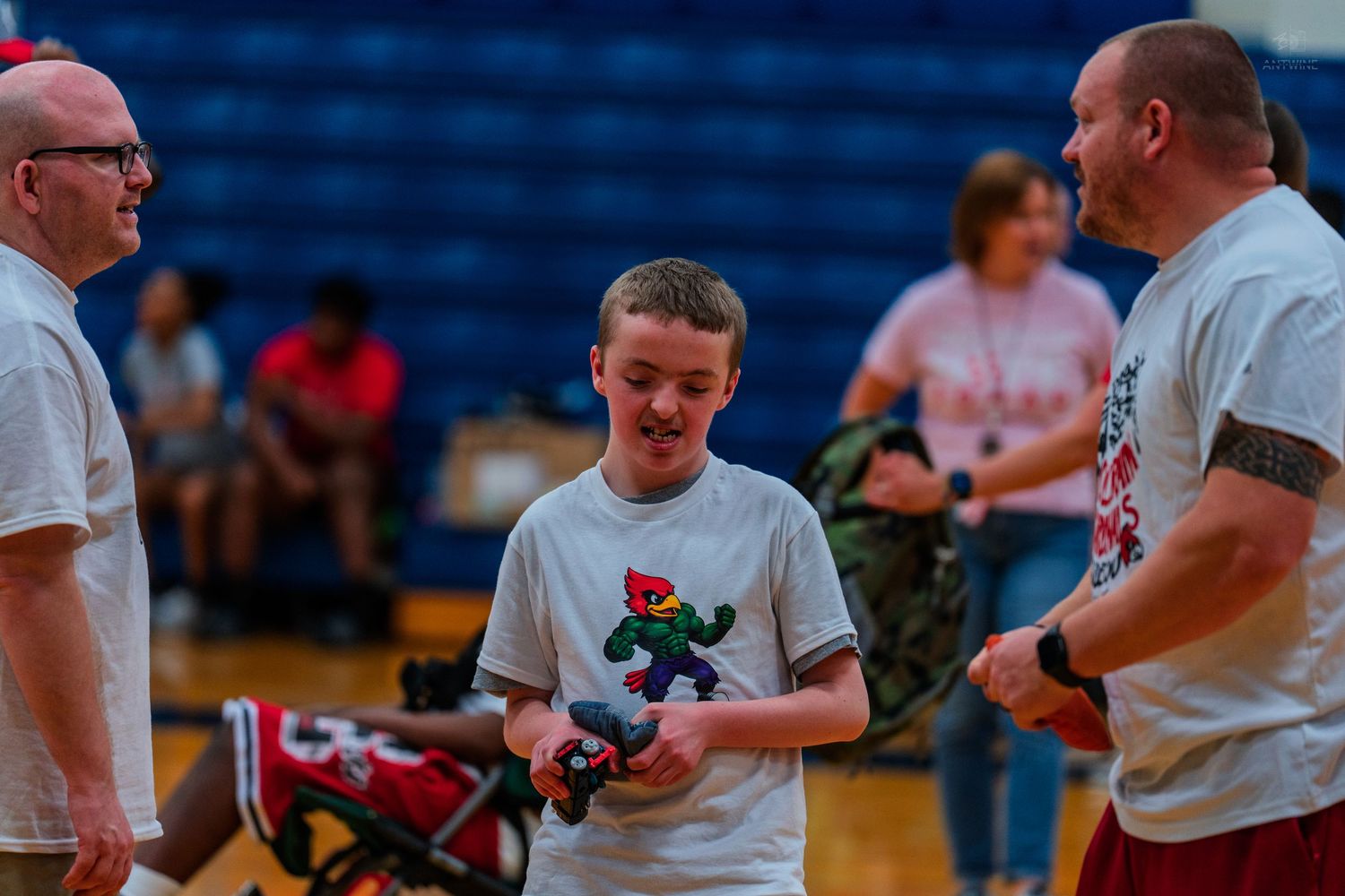 A boy holds a colorful toy while two adults stand nearby in a gymnasium during a school event.