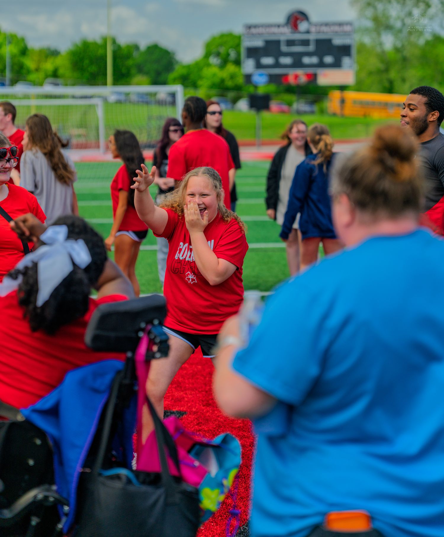 Enthusiastic woman in red shirt gestures excitedly at an outdoor athletic event with spectators and scoreboard.