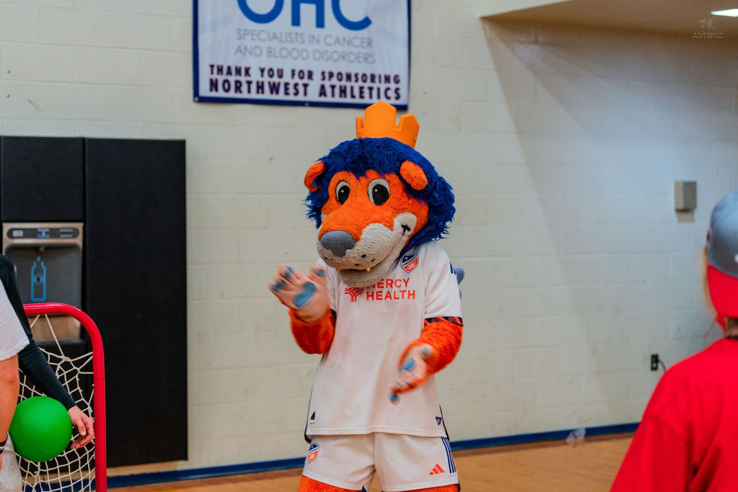 A sports mascot in orange and blue costume waves inside a gymnasium with OHC sponsorship banner.